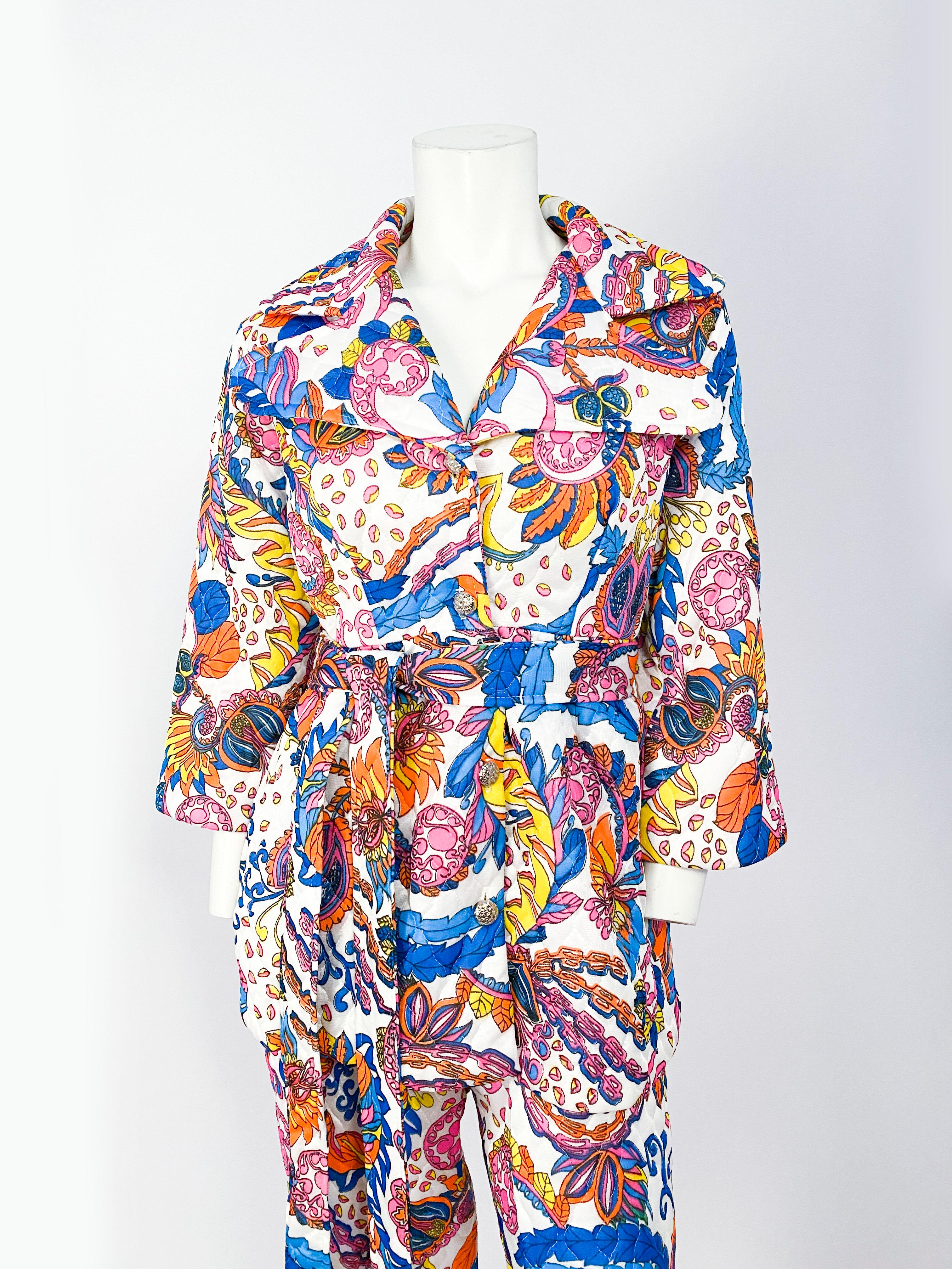 1970s printed polyester pant suit set (jacket, pants, & sash) featuring a psychedelic floral print in neon pinks, orange, blues, and yellows on a white back color. The jacket has a wide flared hem, hidden pockets, metallic buttons, and an enlarged