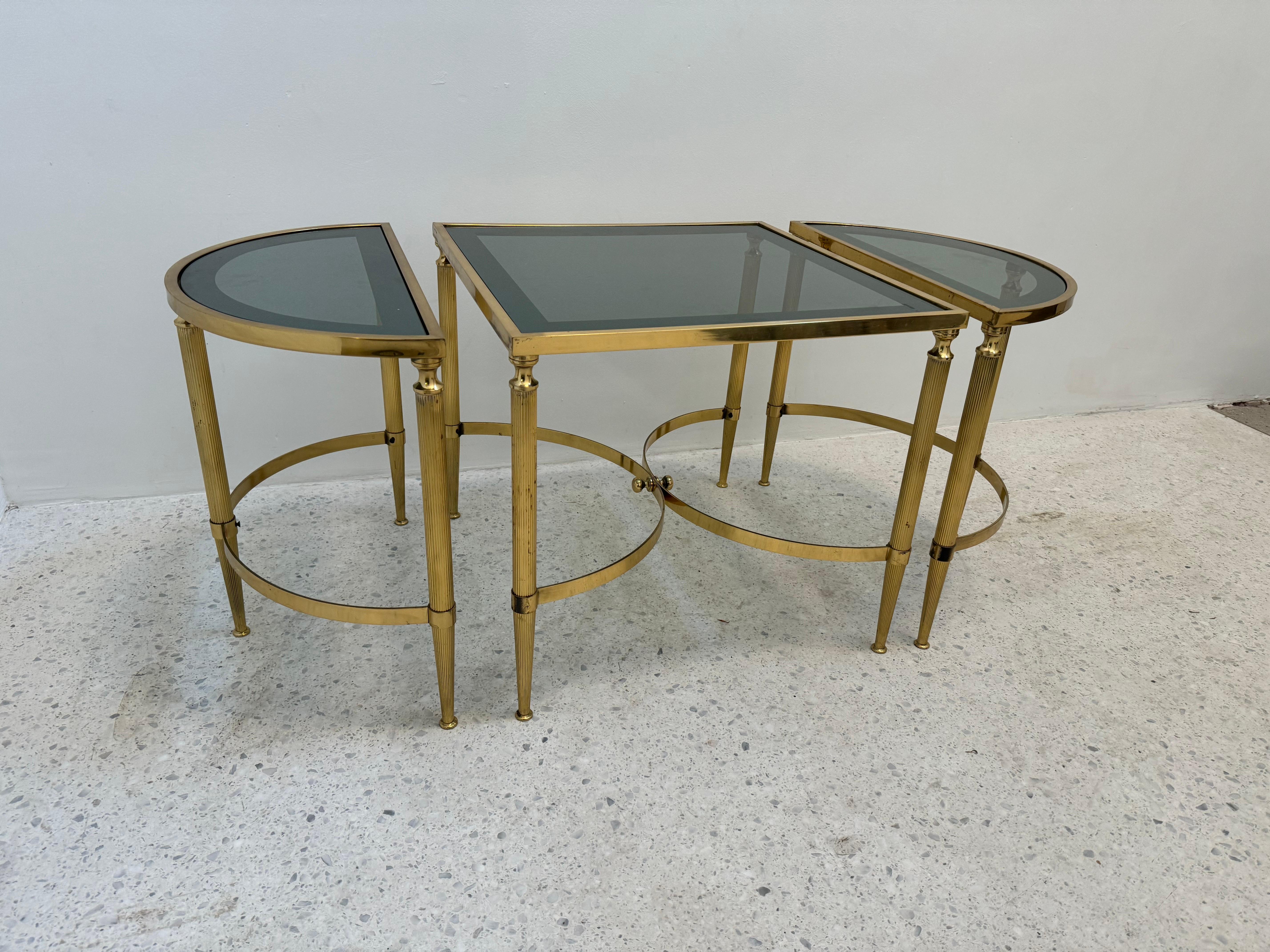 Elegant ensemble de table néoclassique des années 1970 en laiton et verre fumé, composé d'une table centrale carrée flanquée de deux tables d'appoint en demi-lune. Les pieds cannelés en laiton et les brancards circulaires raffinés évoquent le style