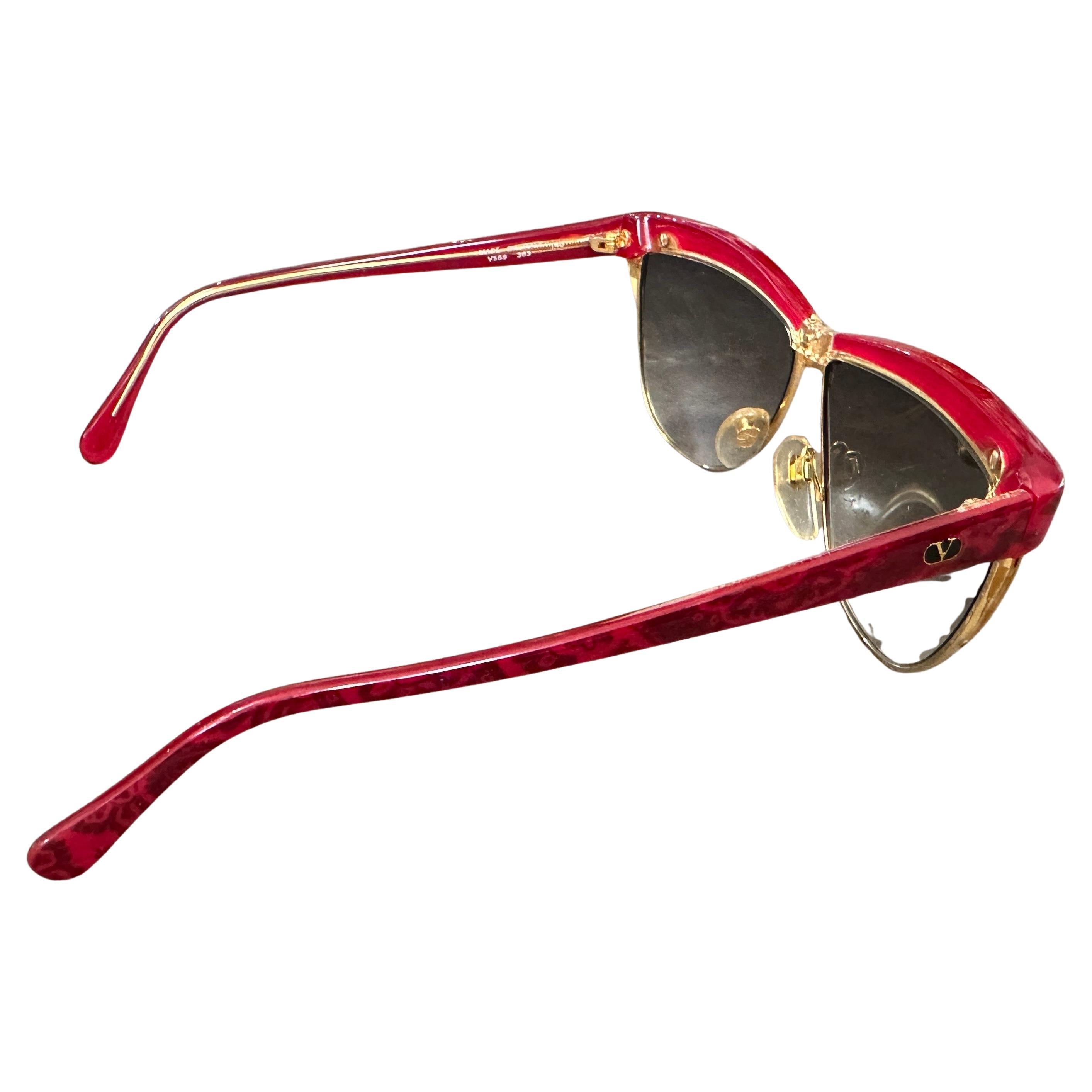 These are a pair of never-used vintage sunglasses by Valentino from the 1970s. They feature an elegant and bold design with a striking red marbled upper frame that extends across the browline, creating a dramatic contrast with the gold-tone metal