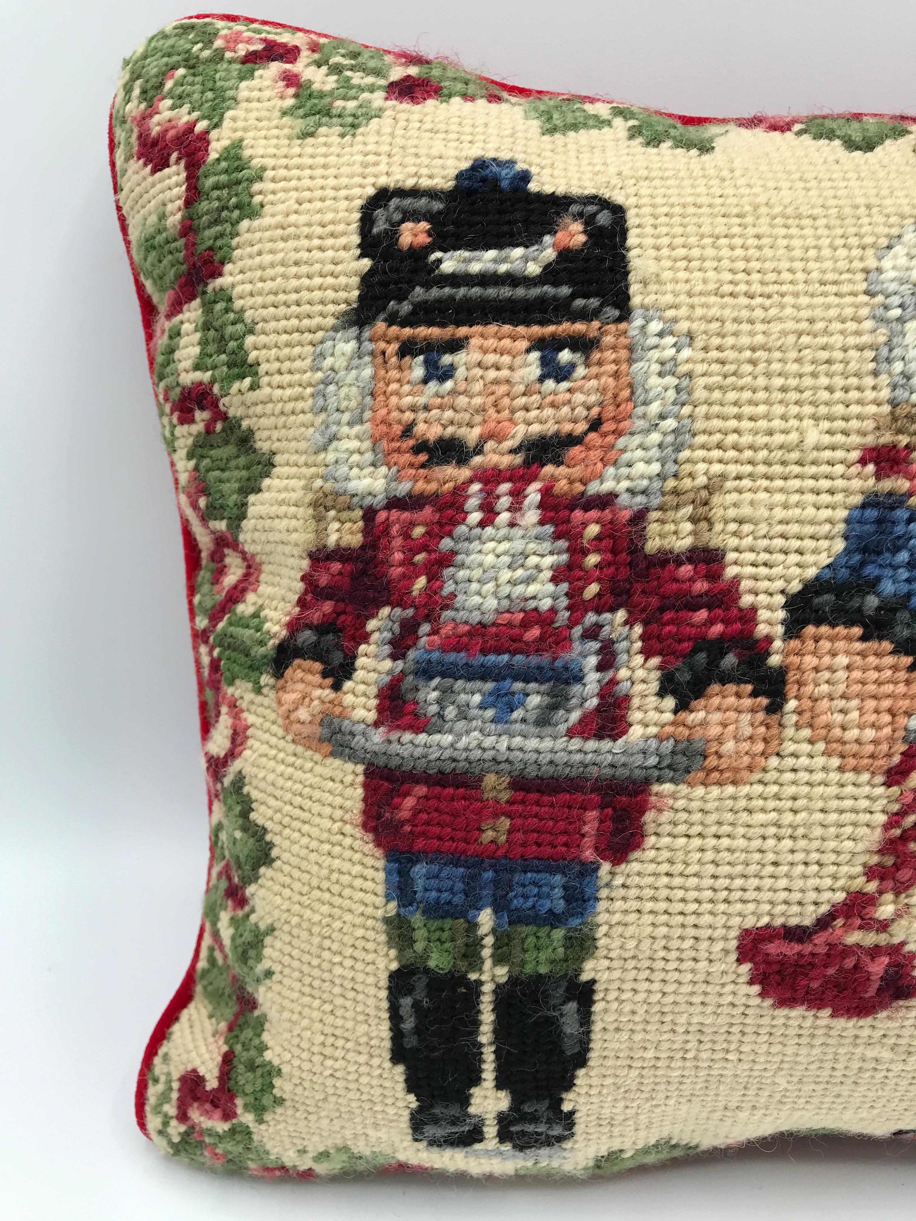 Listed is a festive and seasonal, 1970s wool needlepoint accent pillow. The piece features three nutcrackers on the frontside, with an ivy vine motif along the border. The backside and welt are red velvet, with a hidden zipper closure. Poly-blend