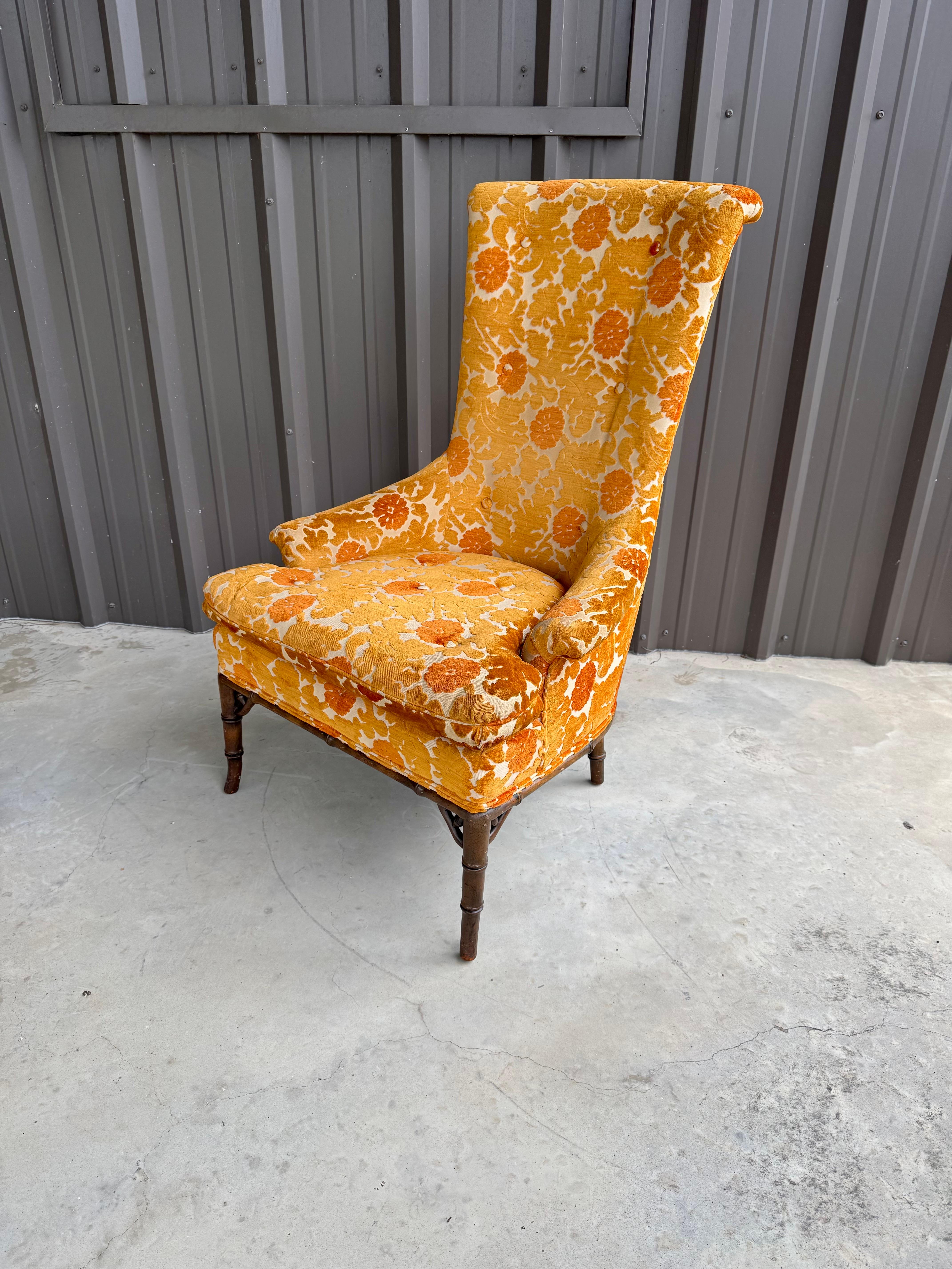 1970s tall-back chair featuring a bold and vibrant orange and gold floral upholstery typical of the era. The chair’s exaggerated high back curves slightly at the top, adding an element of drama and flair. The plush seat cushion and rolled arms give