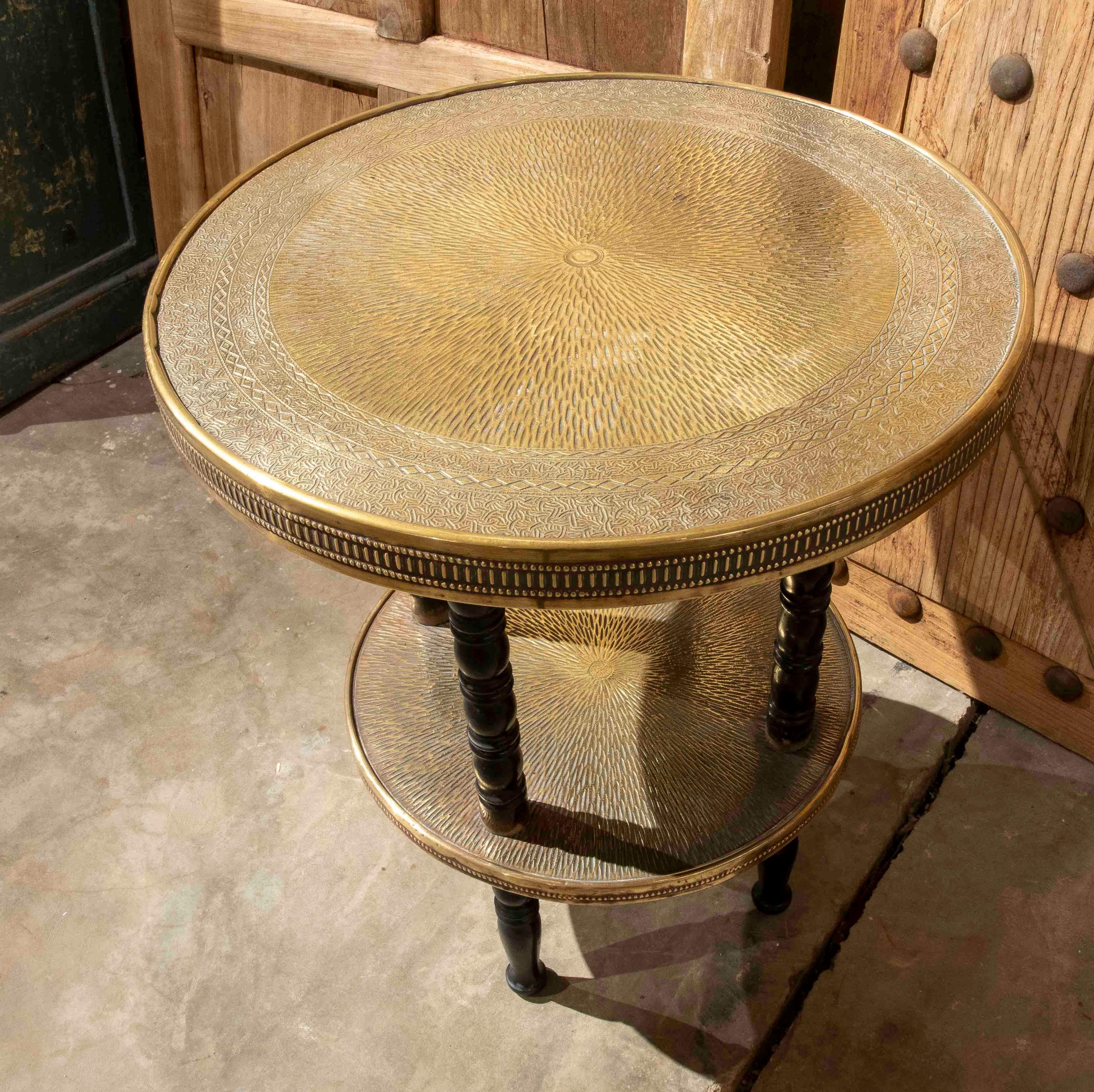 Table d'appoint élégante à deux niveaux avec plateaux circulaires en laiton gaufré et pieds tournés en bois sombre. Le travail artisanal du métal, avec ses gravures géométriques et sa texture martelée, reflète l'influence orientale sur le mobilier