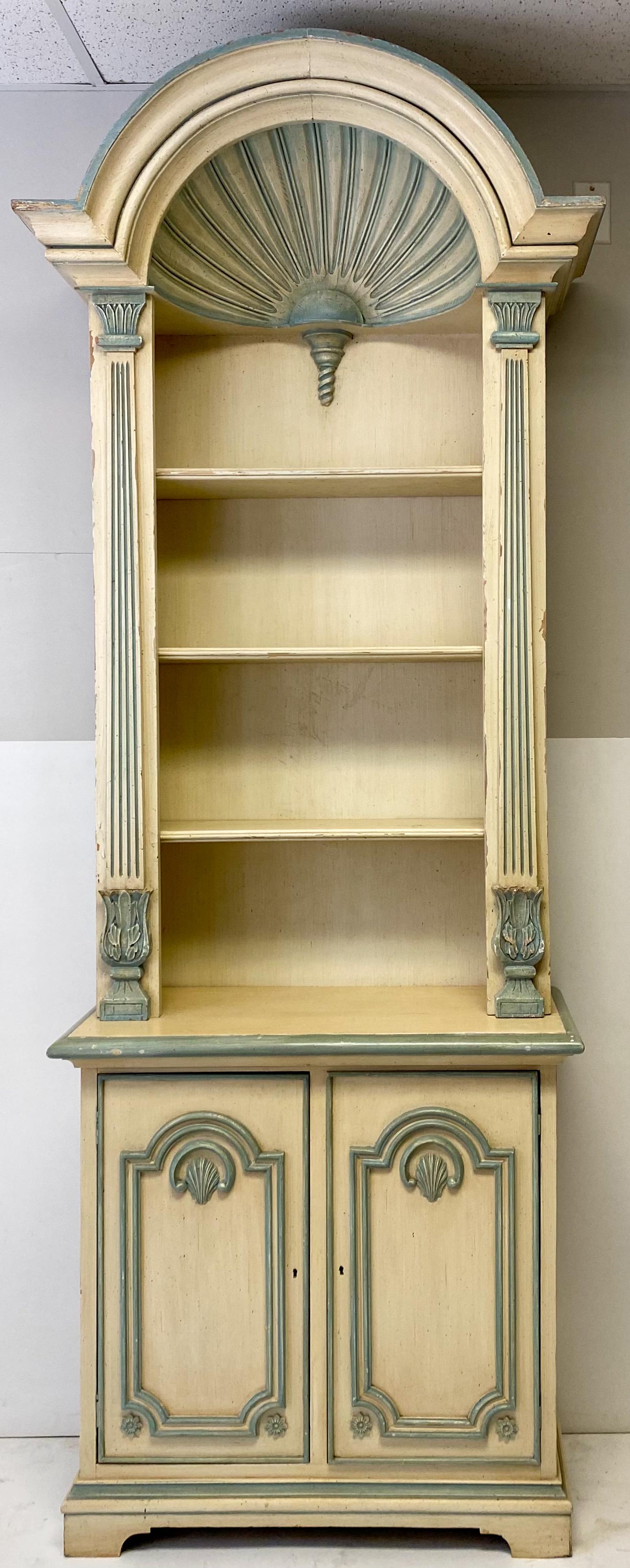 This is a pair of painted grotto inspired carved shell bookcases. They are American and date to the later part of the 20th century. They are unmarked and there is wear to the ivory and blue painted finish.