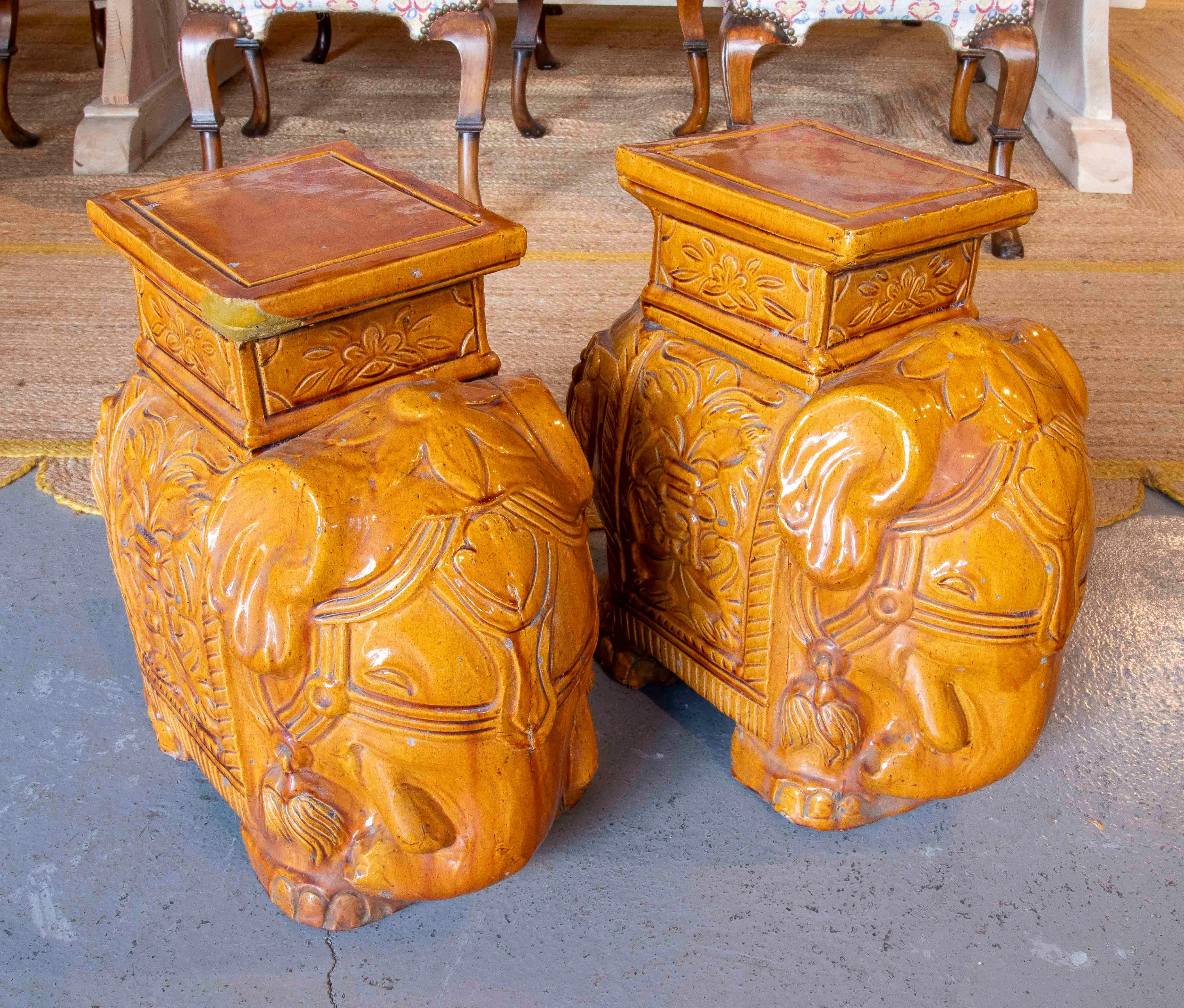 Decorative pair of glazed ceramic stools or stands in a warm mustard hue, modeled in the shape of elephants richly adorned with floral and ornamental motifs. The flat, square top allows them to be used as auxiliary seats, side tables, or pedestals