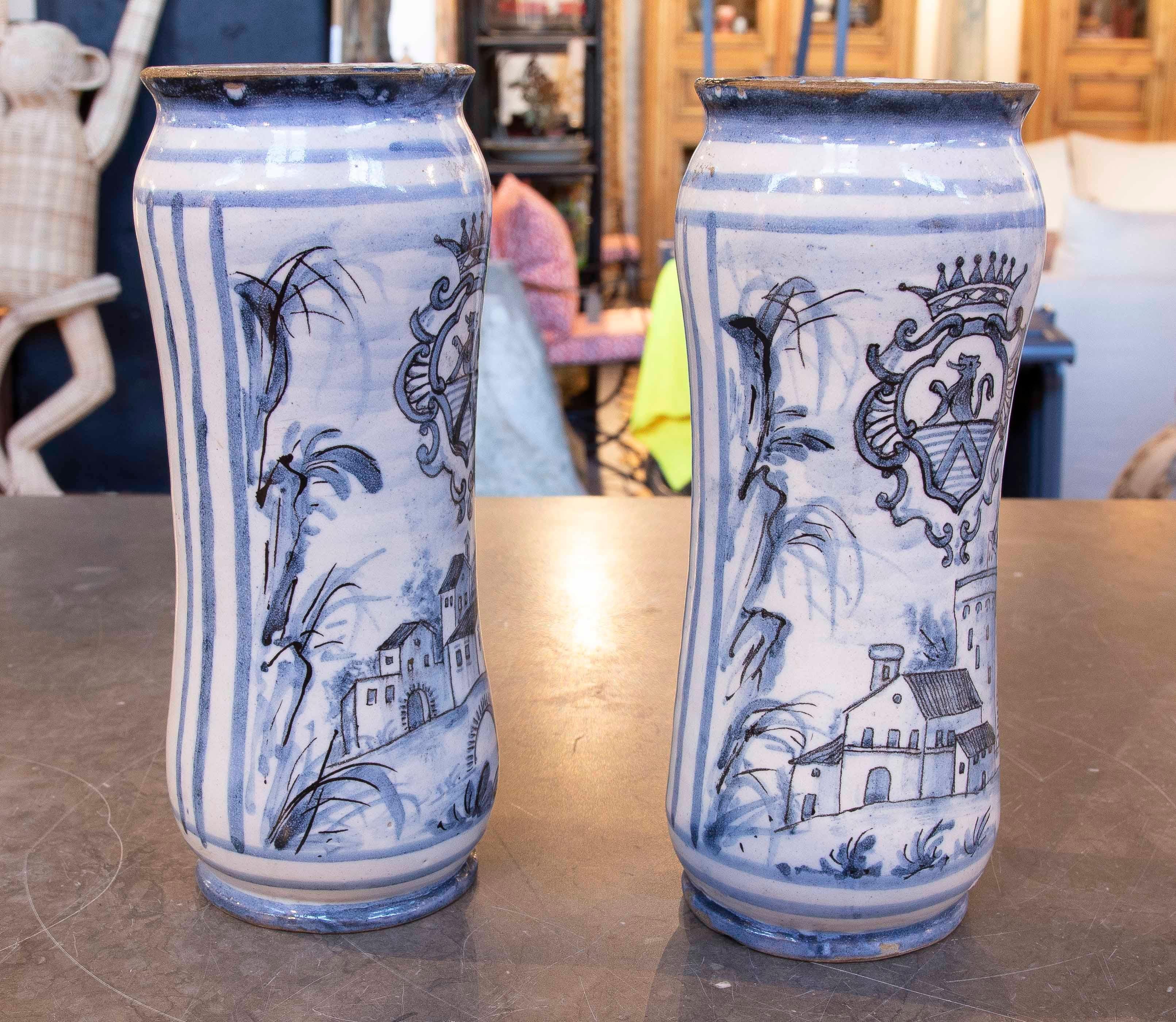 Ensemble de deux vases en céramique émaillée, décorés de motifs héraldiques et de paysages ruraux, peints à la main dans des tons bleus. Chaque pièce présente un blason avec couronne, accompagné de scènes évoquant l'architecture traditionnelle et la