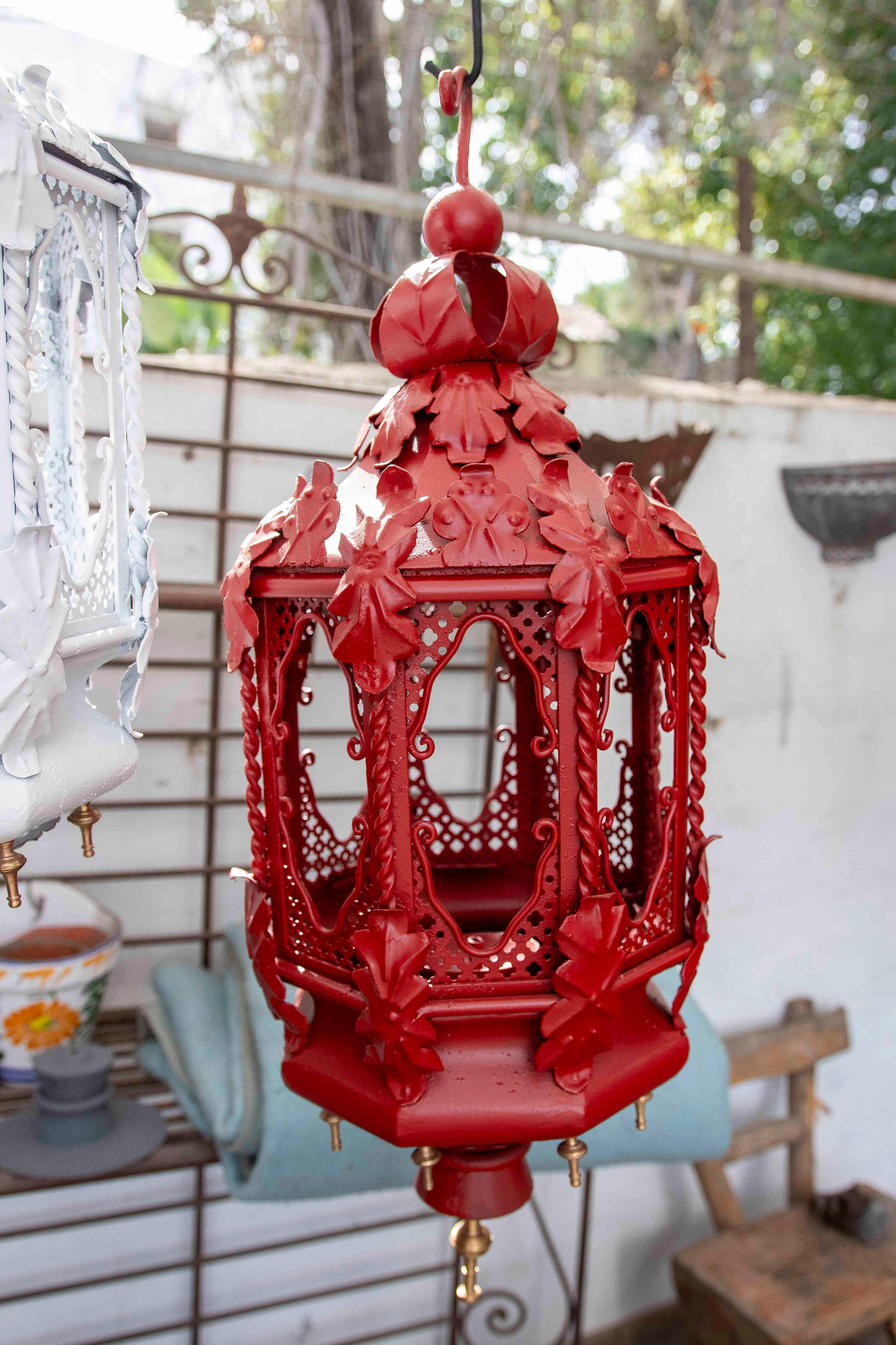 Elegante coppia di lampade a sospensione in ferro battuto di colore rosso vivo, ornate con dettagli ornamentali di foglie e motivi traforati che evocano uno stile vintage e artigianale. La sua struttura esagonale con finestre decorate e le raffinate