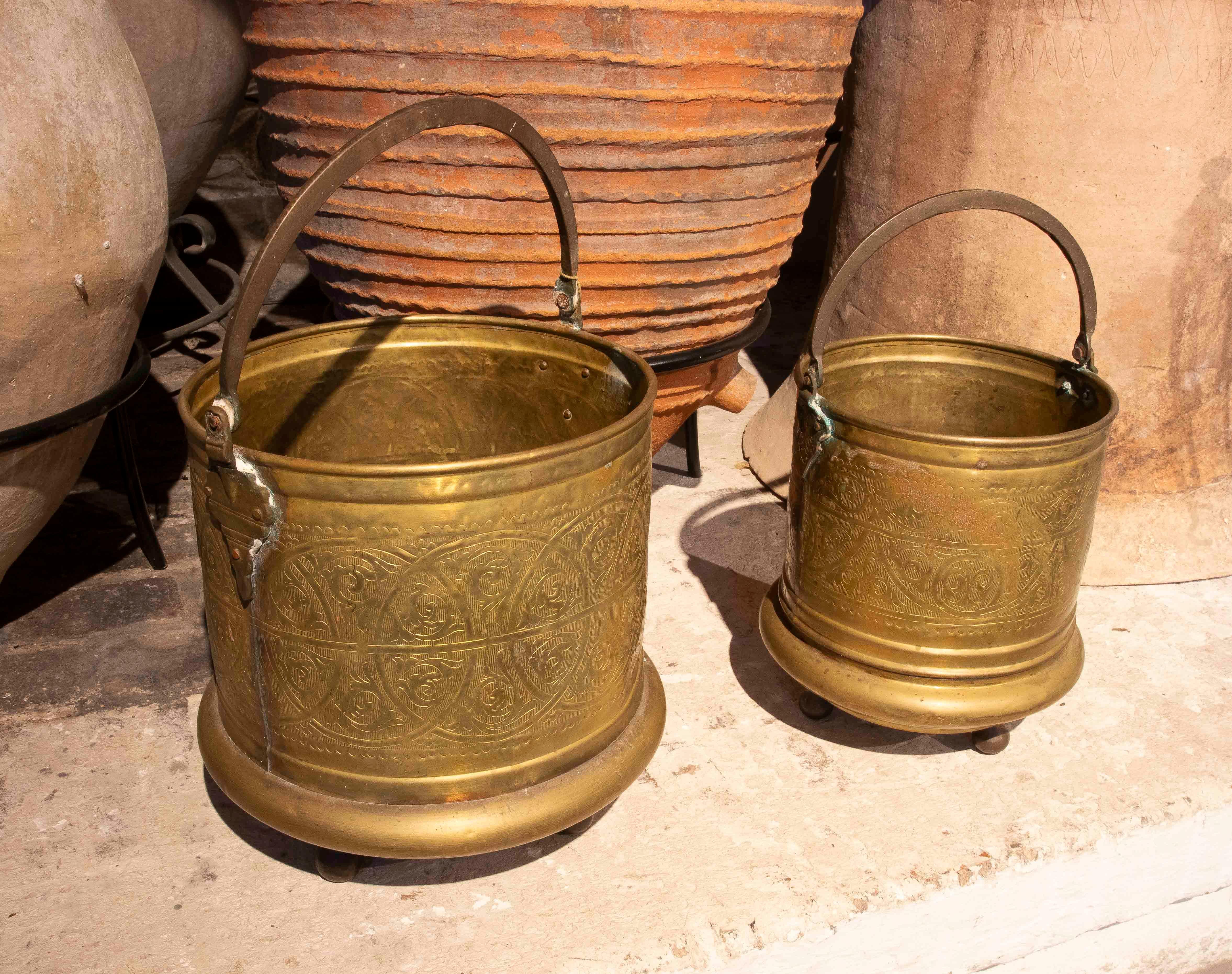 Une élégante paire de seaux décoratifs en laiton, finement estampés de motifs géométriques et floraux répétés dans des frises circulaires. Chaque seau est doté d'une poignée en fer forgé et d'une base moulée avec de petits pieds sphériques, ce qui