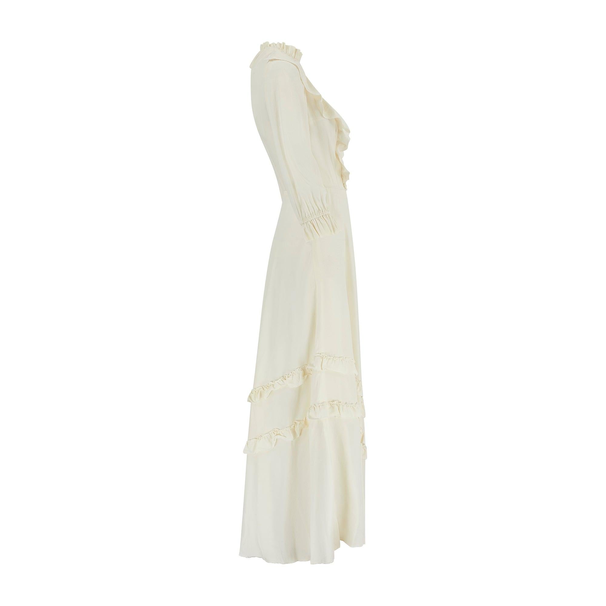 This 1970 maxi dress encompasses a bohemian aesthetic and is crafted from cream silk or a silk - rayon blend.  It has elements of Victoriana in the high, ruffle neckline and formal, long silhouette. The sleeves are long with pretty ruffle cuffs. The