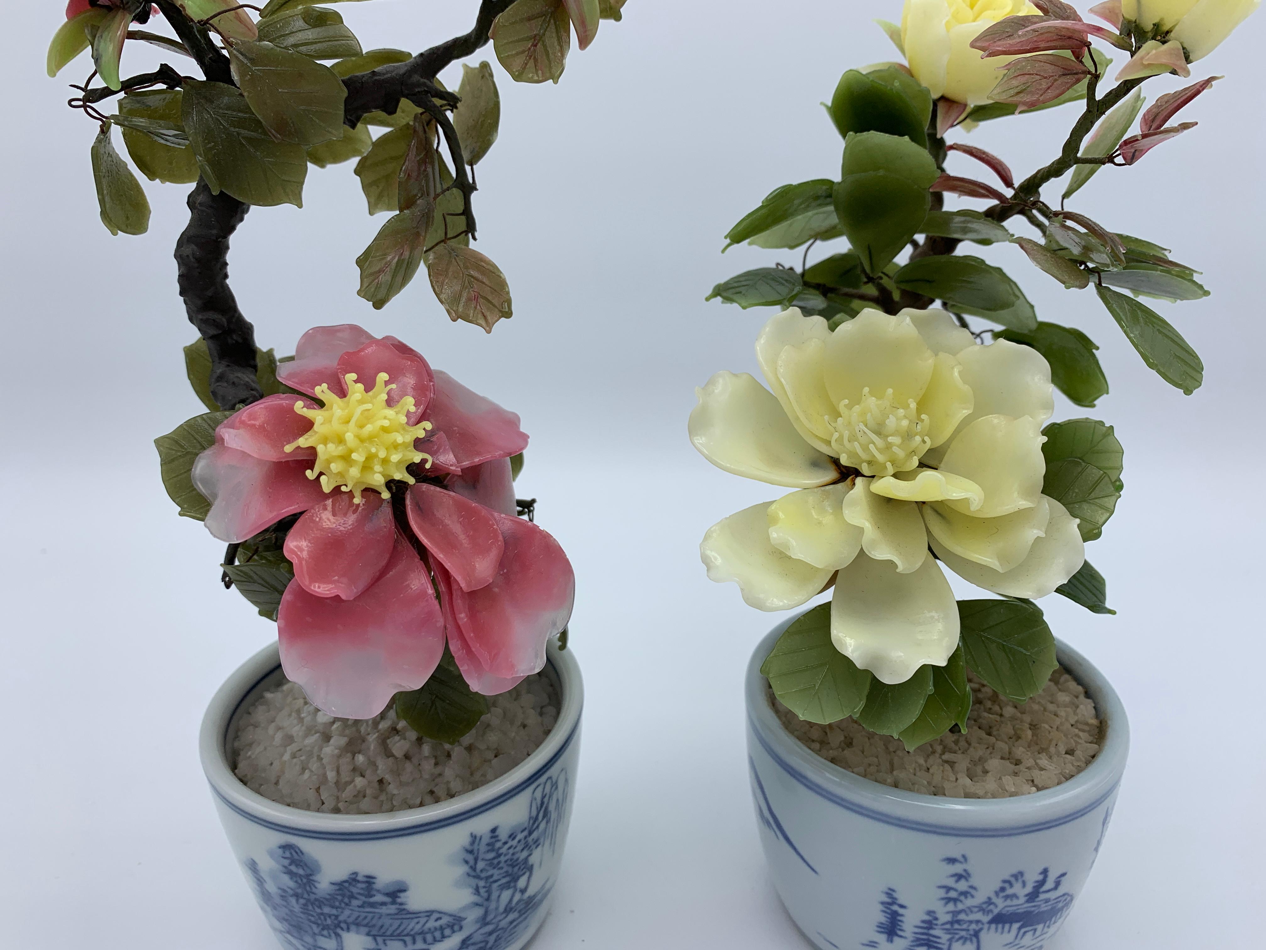 Offered is a fabulous, pair of 1970s jade tree sculptures in the form of peonies. The pair includes one pink with yellow peony and one solid-yellow peony. The pair are each 'planted' in blue and white cachepots, with ornate chinoiserie scenery