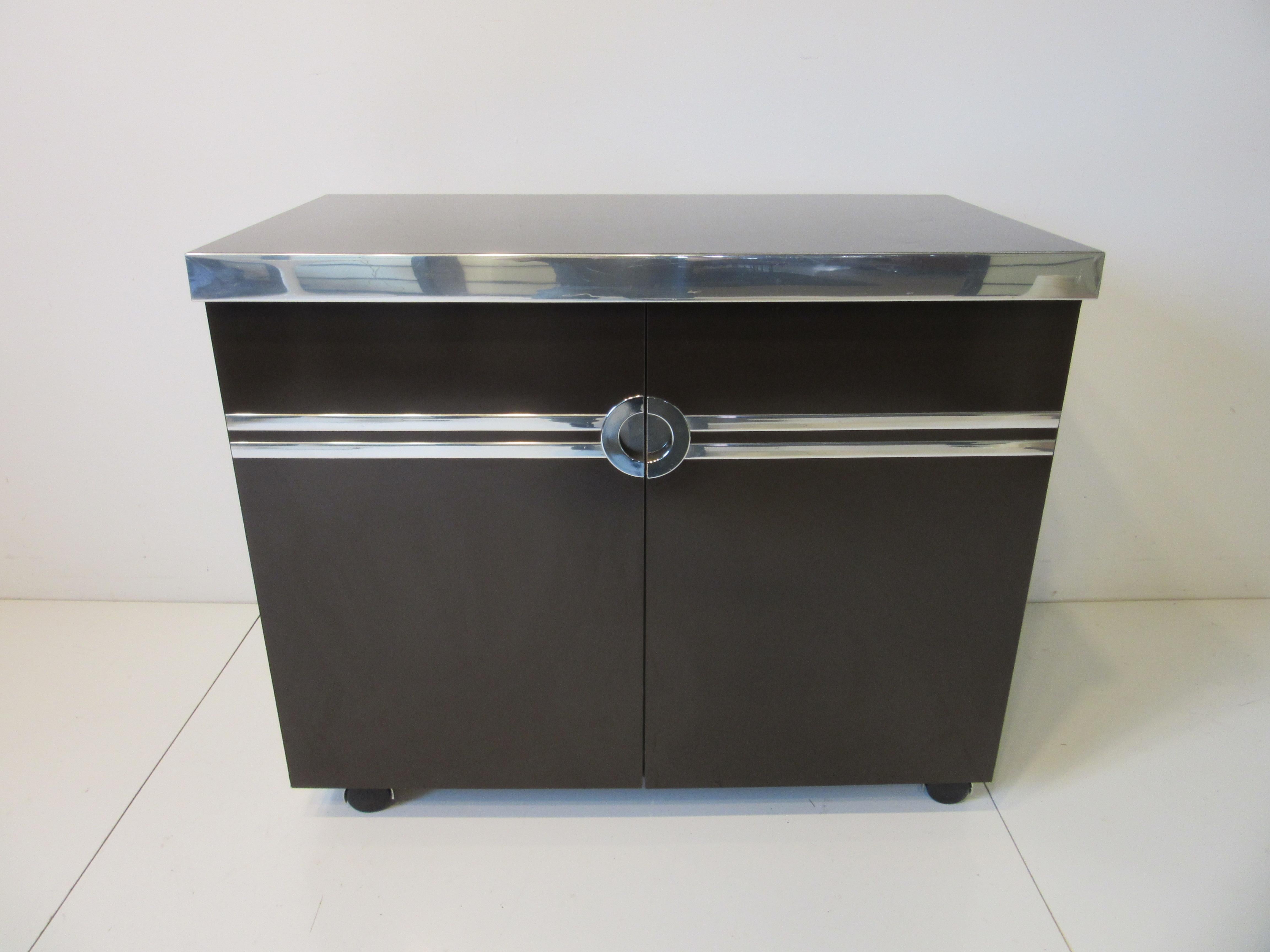 A very well crafted rich chocolate brown rolling bar cart with flip up top reveling a mirrored back , built in ice bucket and areas for glasses and bottle storage in a brushed stainless laminate . The bar has polished aluminum trim to the edges ,