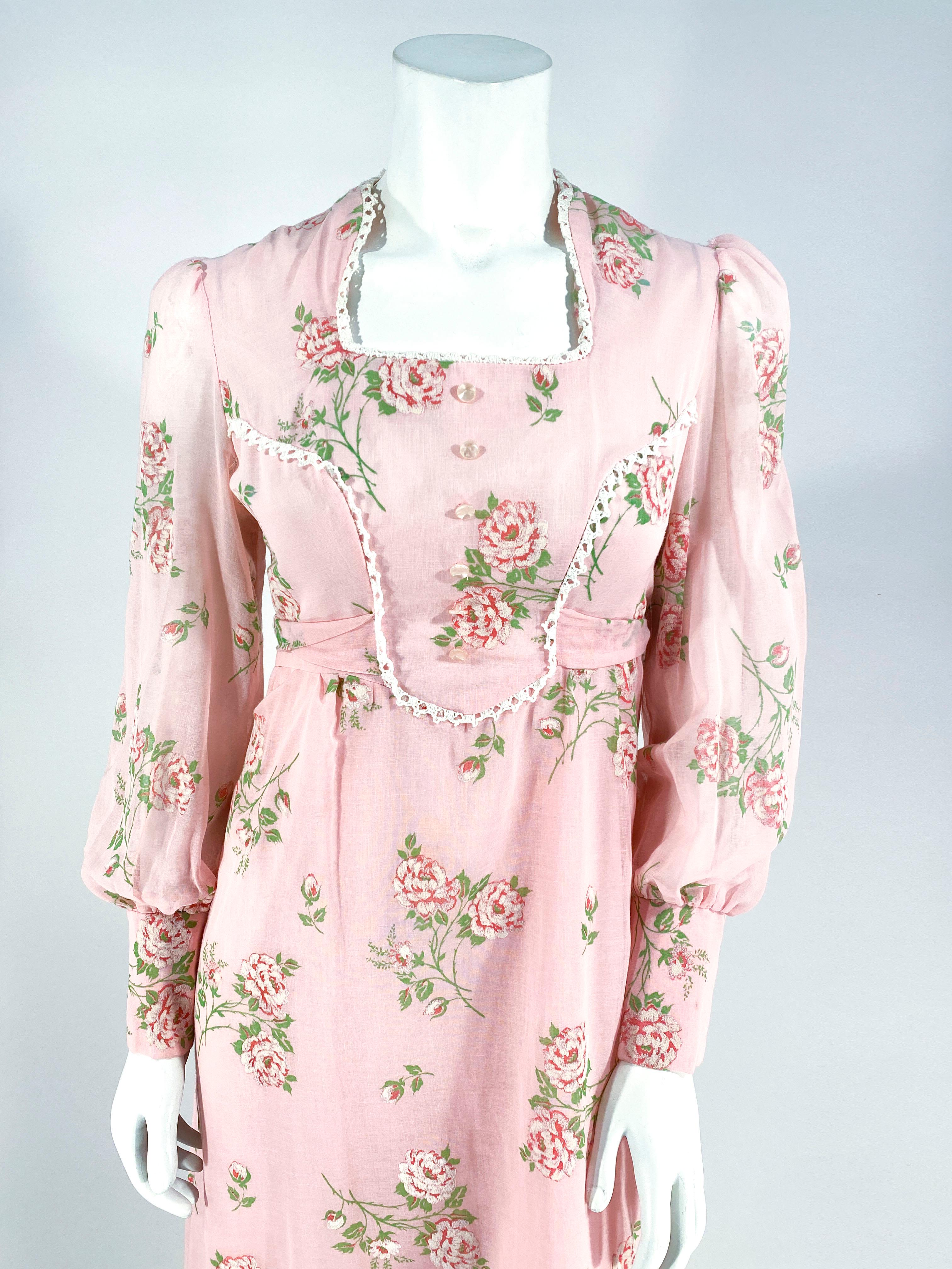 Vestido campero de gasa de algodón rosa de los años 70, estampado con rosas rosas y verdes flocadas para darle textura. En  El escote cuadrado y el corpiño de corte princesa están rematados con encaje de ganchillo y un fajín doble aplicado que se