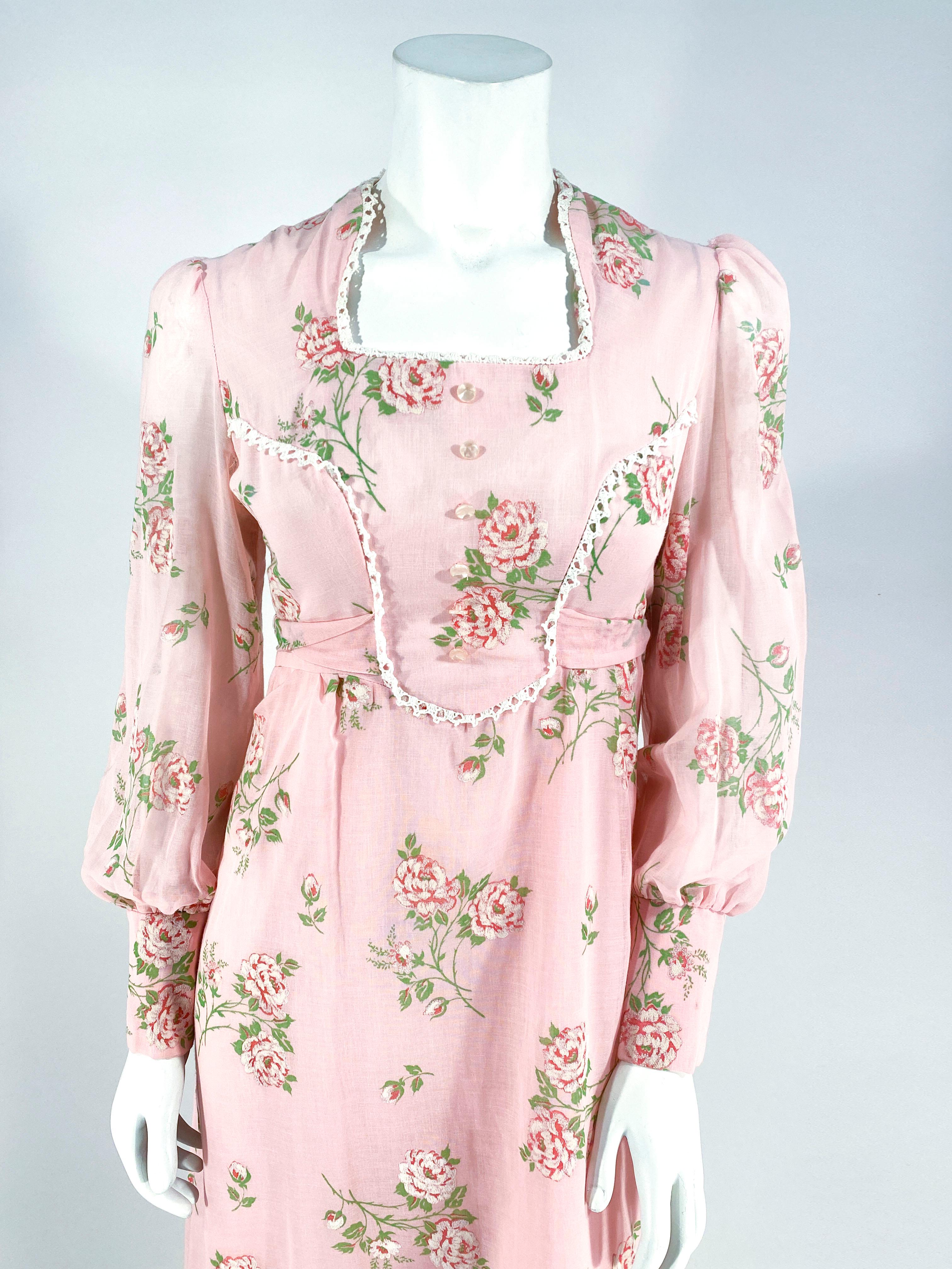 1970s Pink cotton voile cottage dress printed with pink and green roses flocked for texture. The  square neckline and princess cut bodice is finished with crochet lace and an applied double sash that ties around the back. The body of the dress if