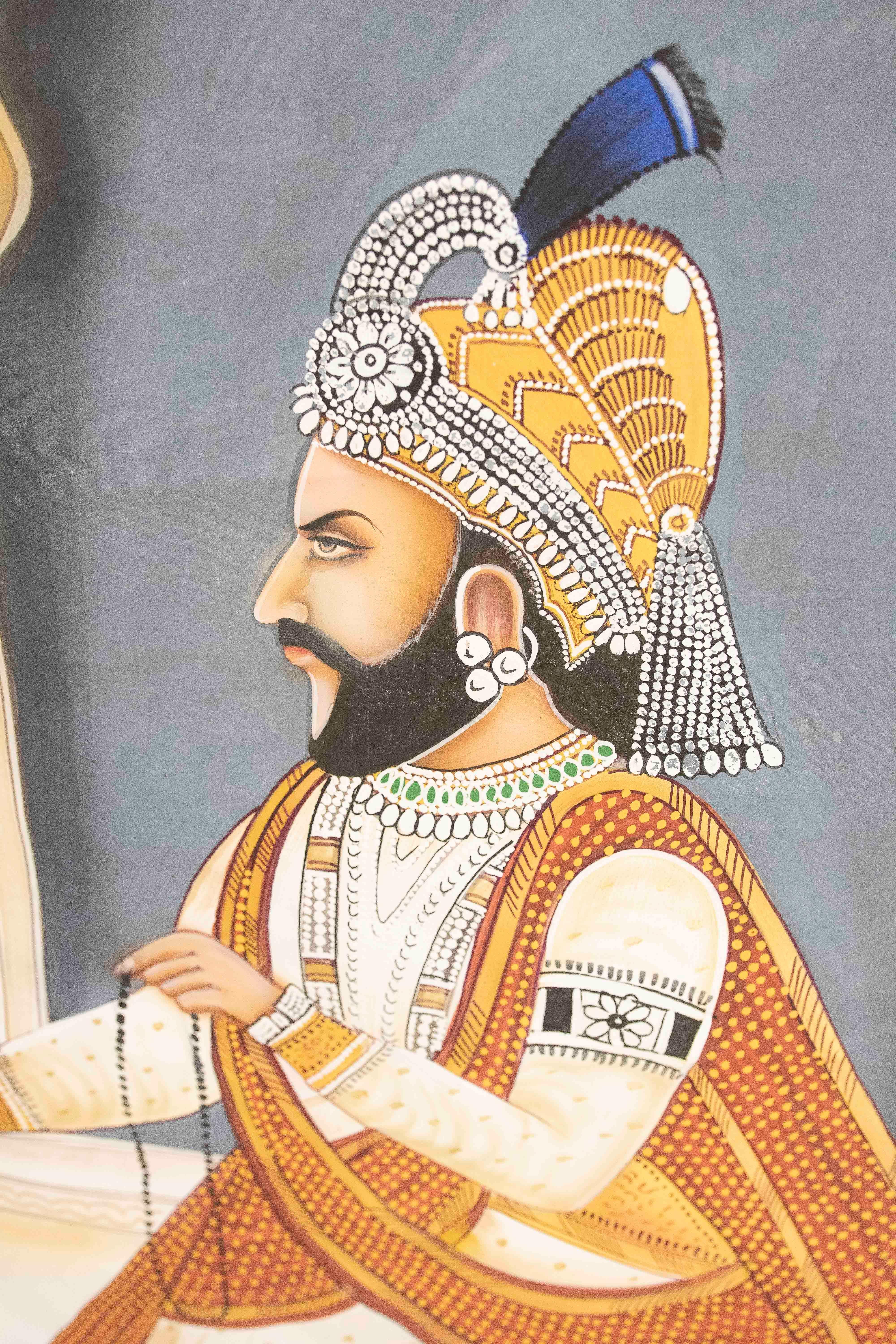 Esta imponente obra de estilo miniaturista rajastaní representa a un guerrero real indio en una escena ceremonial. La figura, de perfil, aparece vestida con una túnica blanca con bordes dorados, una capa roja bordada con finos detalles y un regio