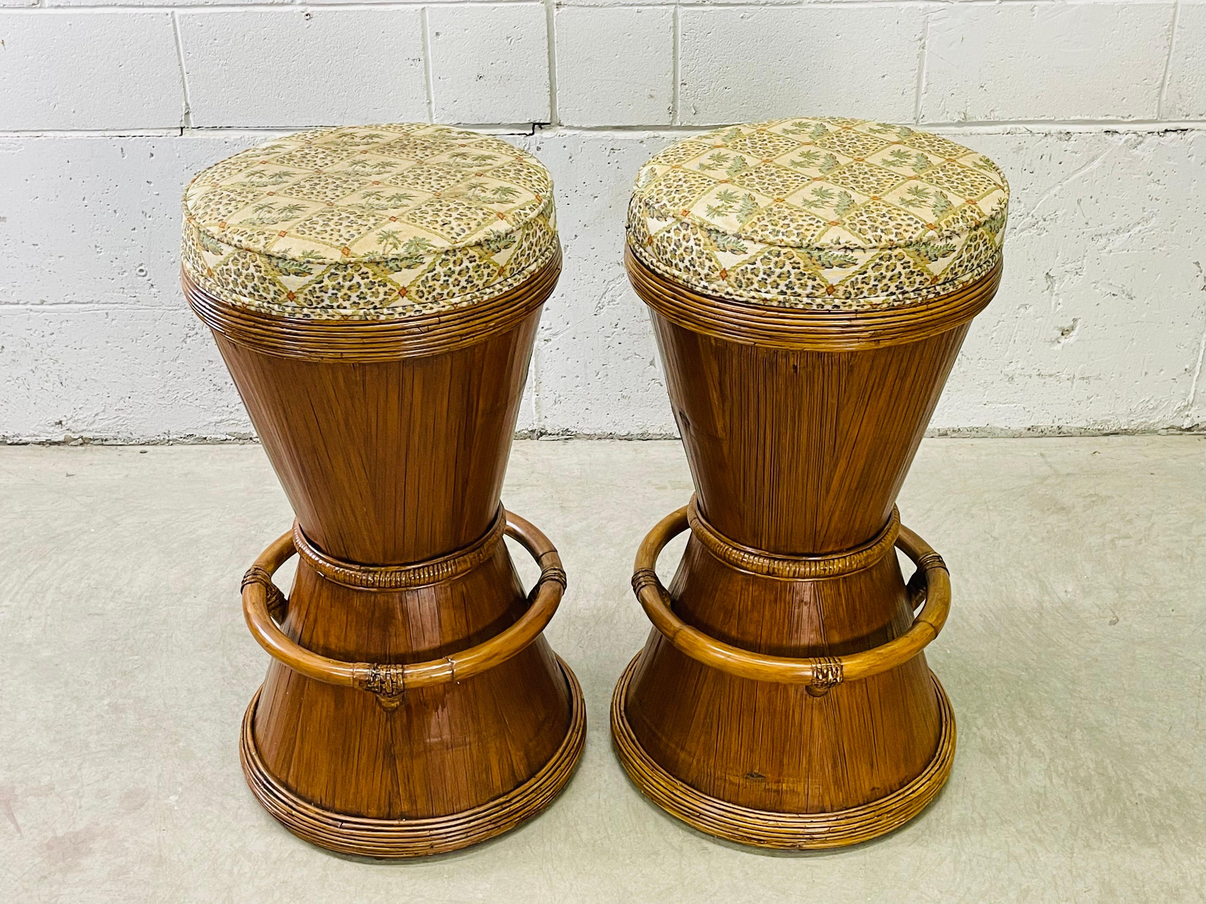 Vintage 1970s pair of tall round drum style rattan bar stools with original fabric seats. The stools have a round accented foot rest which is 11.5” H. No marks.