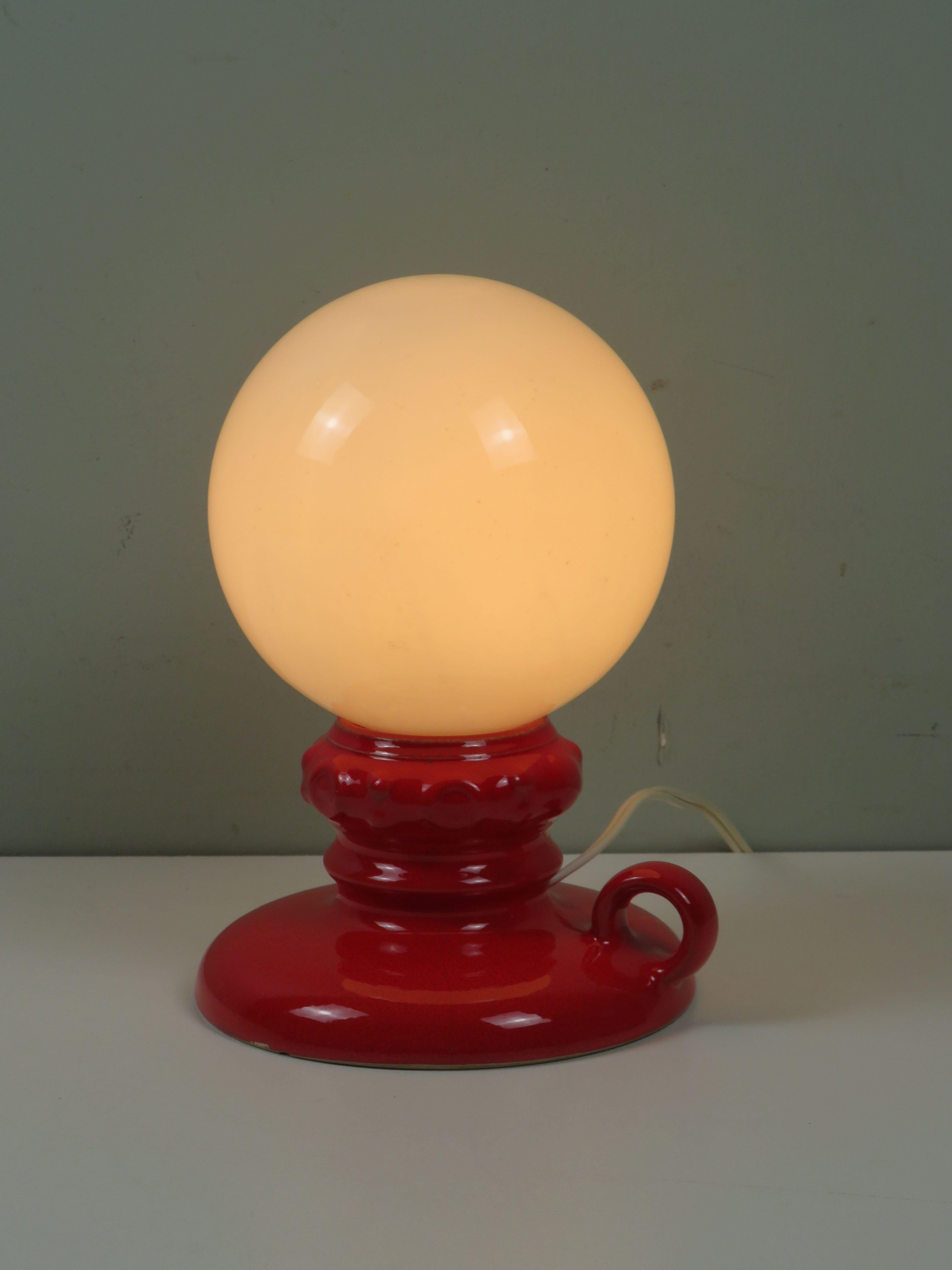 Une lampe de table vibrante et rare, conçue et produite par Massive Belgium dans les années 1970. Cette pièce se compose d'une base en céramique rouge brillante aux contours ornés et d'une poignée circulaire, surmontée d'un globe classique en verre
