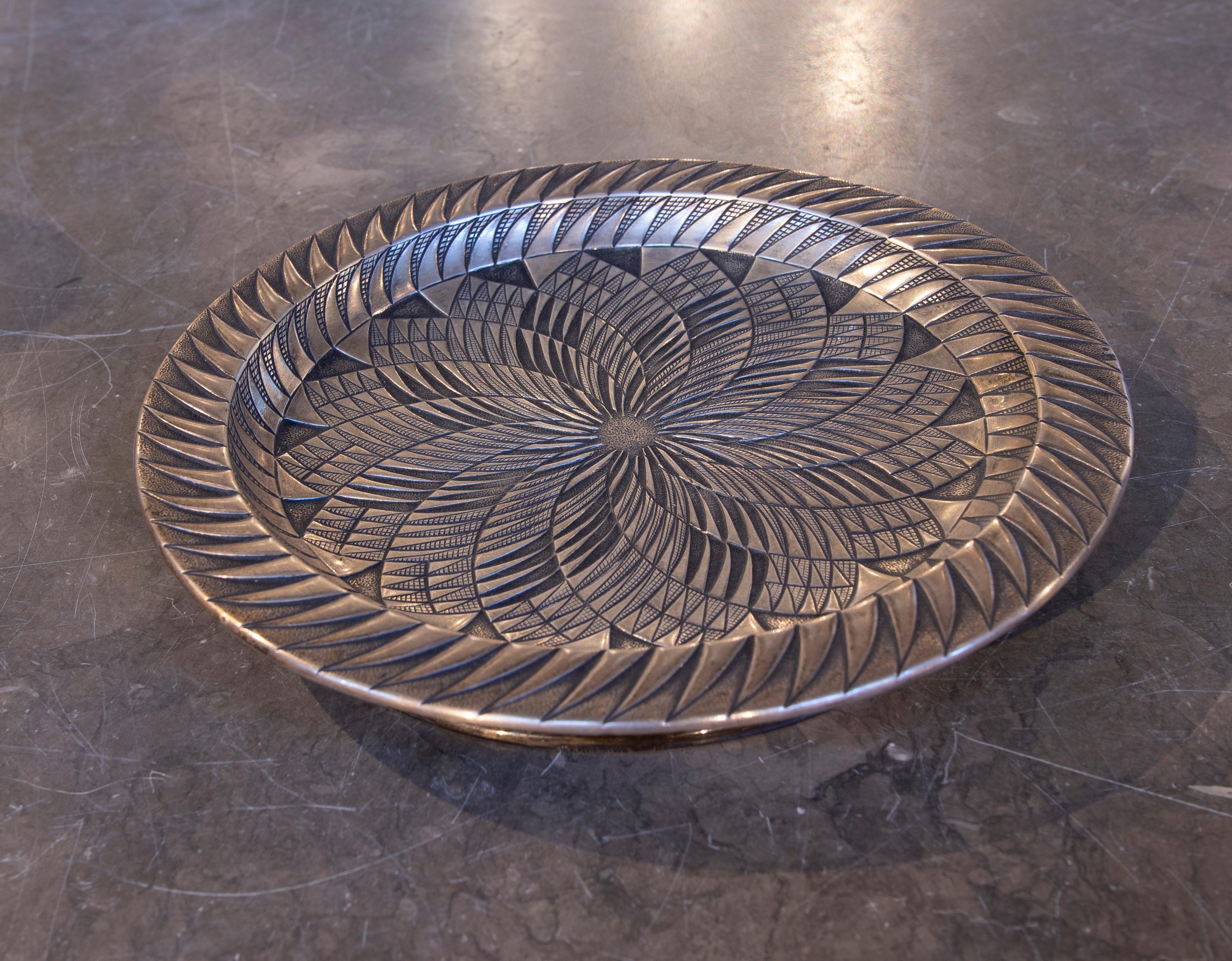 Superbe assiette décorative en argent avec un design gravé présentant des motifs géométriques qui convergent pour un effet visuel élégant. Sa finition sombre et ses détails symétriques lui confèrent un style sophistiqué et intemporel, idéal pour
