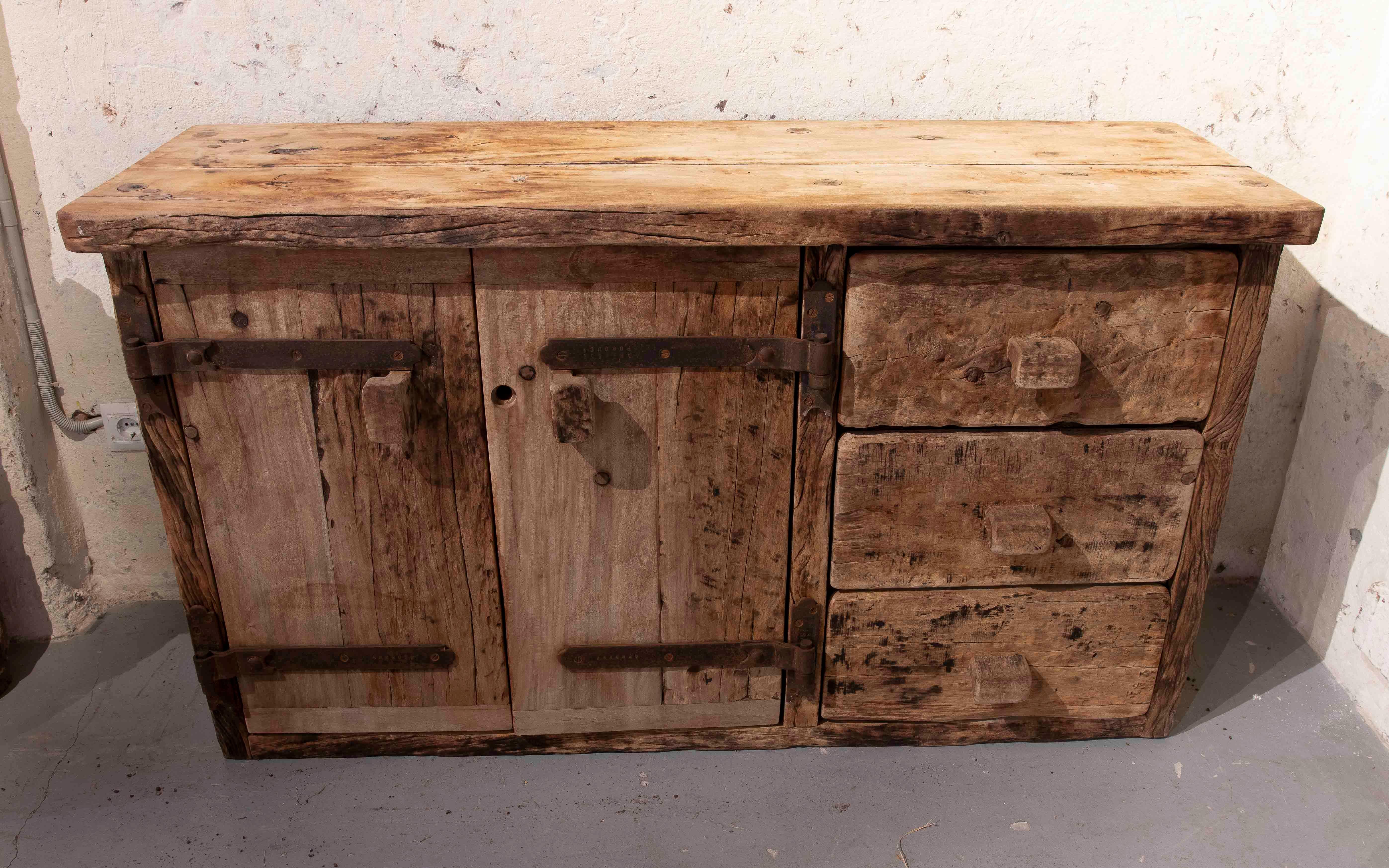 Imposing solid wood sideboard with a rustic and aged finish that highlights its authentic and artisanal character. Its robust design includes two doors with wrought iron hardware and three side drawers with hand-carved handles, providing a natural