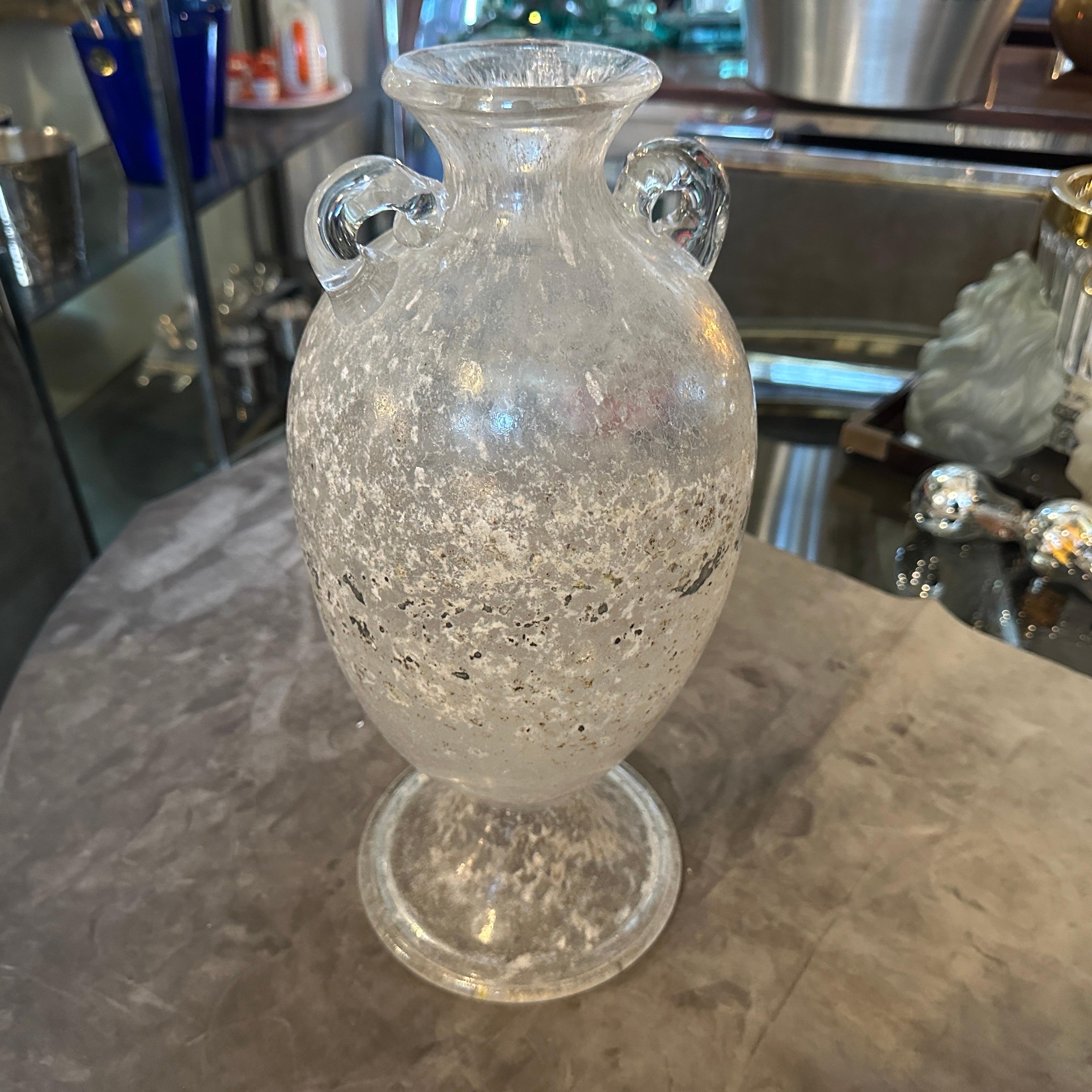 Questo è un vaso ad anfora in vetro di Murano Scavo degli anni '70, attribuito a Seguso, una rinomata famiglia di vetrai di Murano. Il vaso presenta la tecnica dello Scavo, che gli conferisce un aspetto antico e rovinato dalle intemperie, simile ai