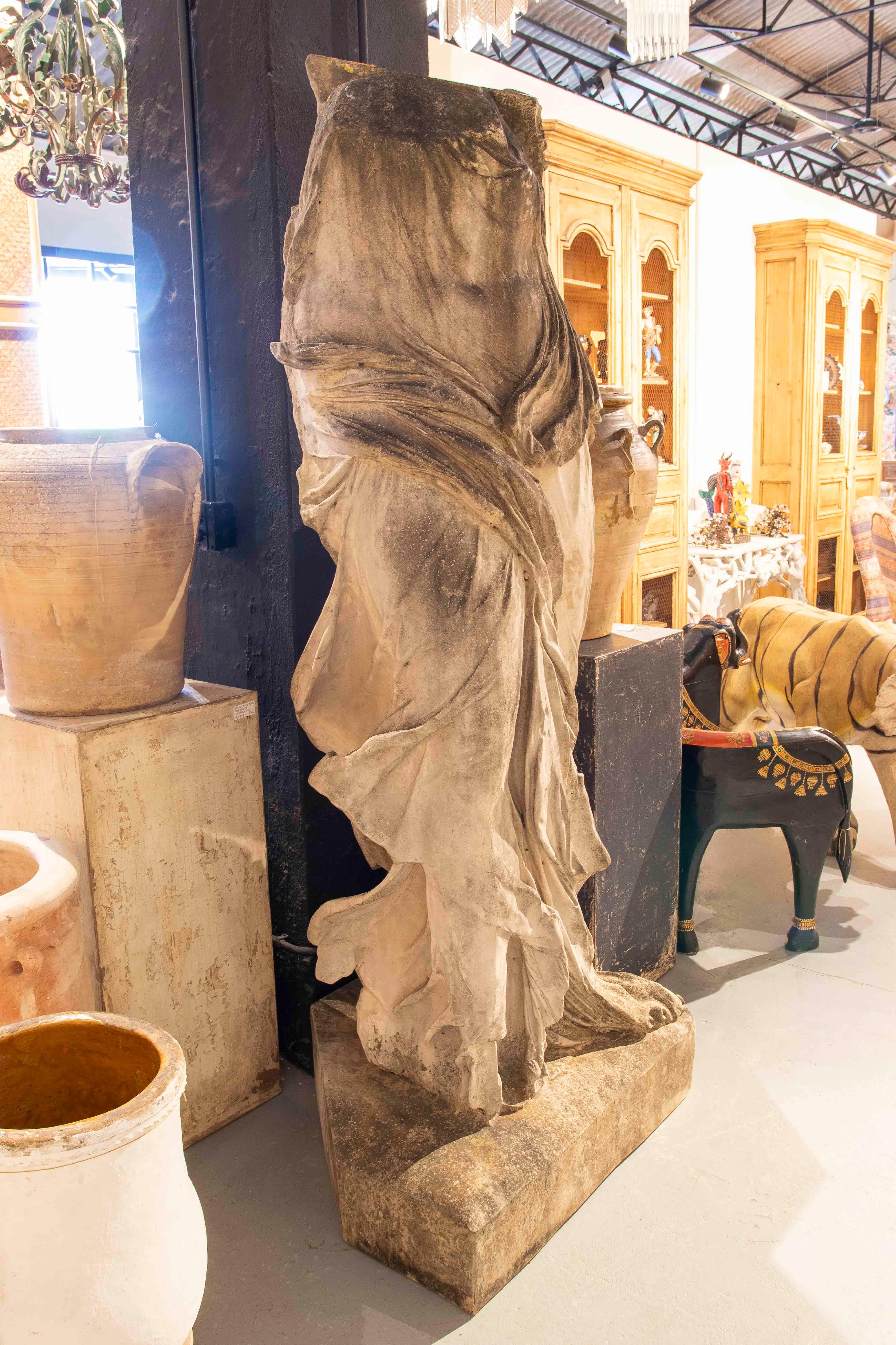 Imponente fragmento de una monumental escultura clásica de resina que representa a la Victoria de Samotracia envuelta en un dinámico y delicado drapeado. La pieza destaca por su drapeado, que sugiere movimiento y fluidez, demostrando una gran