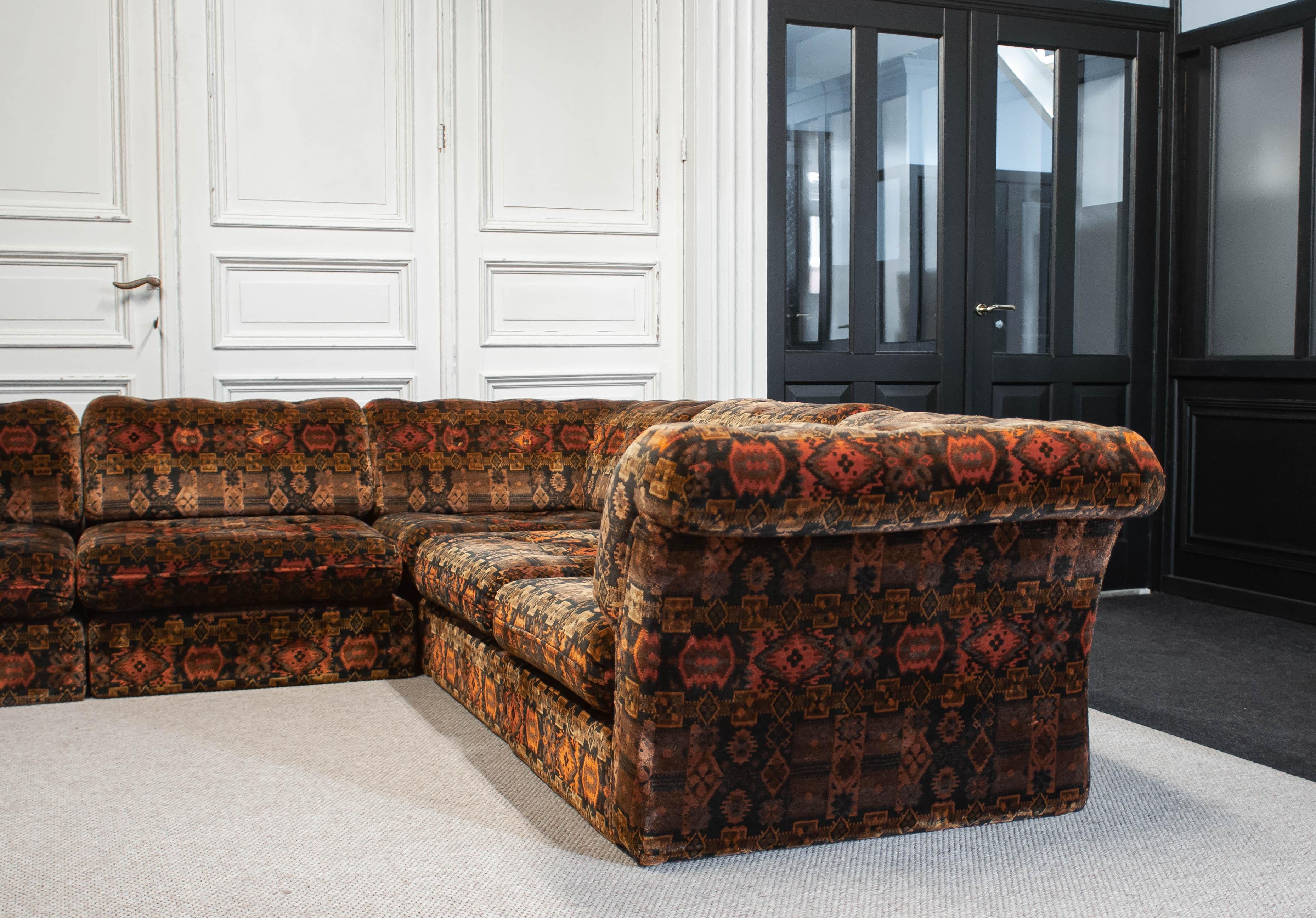 A striking 1970s modular sofa composed of seven individual sections, upholstered in a richly textured velour fabric. The color palette blends deep browns with warm oranges, touches of golden hues, and subtle blacks, creating a bold, almost