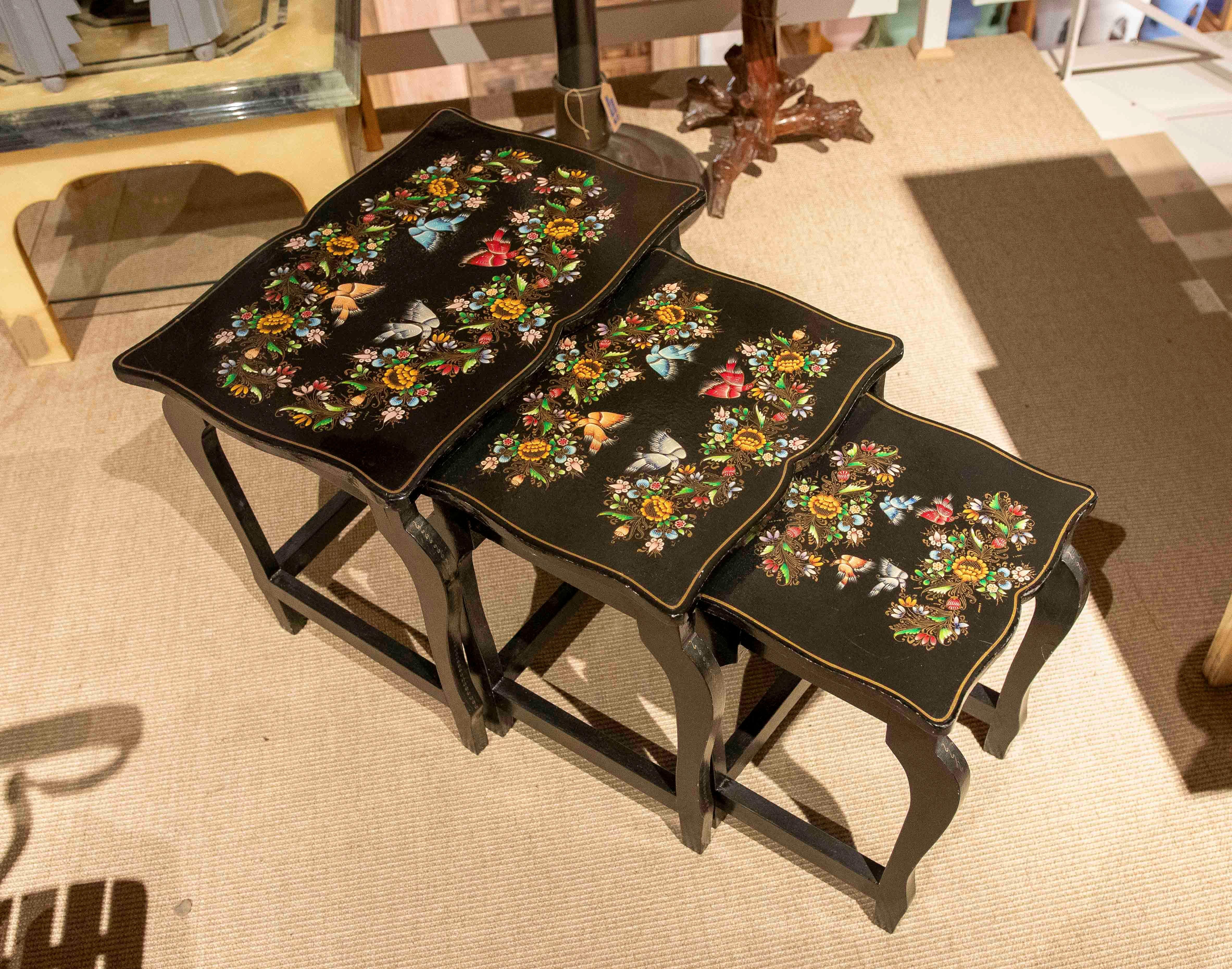 Set di tavolini in legno con finitura nera e delicate decorazioni floreali dipinte a mano in colori vivaci. Ogni tavolo presenta un design elegante con bordi curvi e gambe stilizzate, che evocano uno stile vintage con influenze classiche. La sua