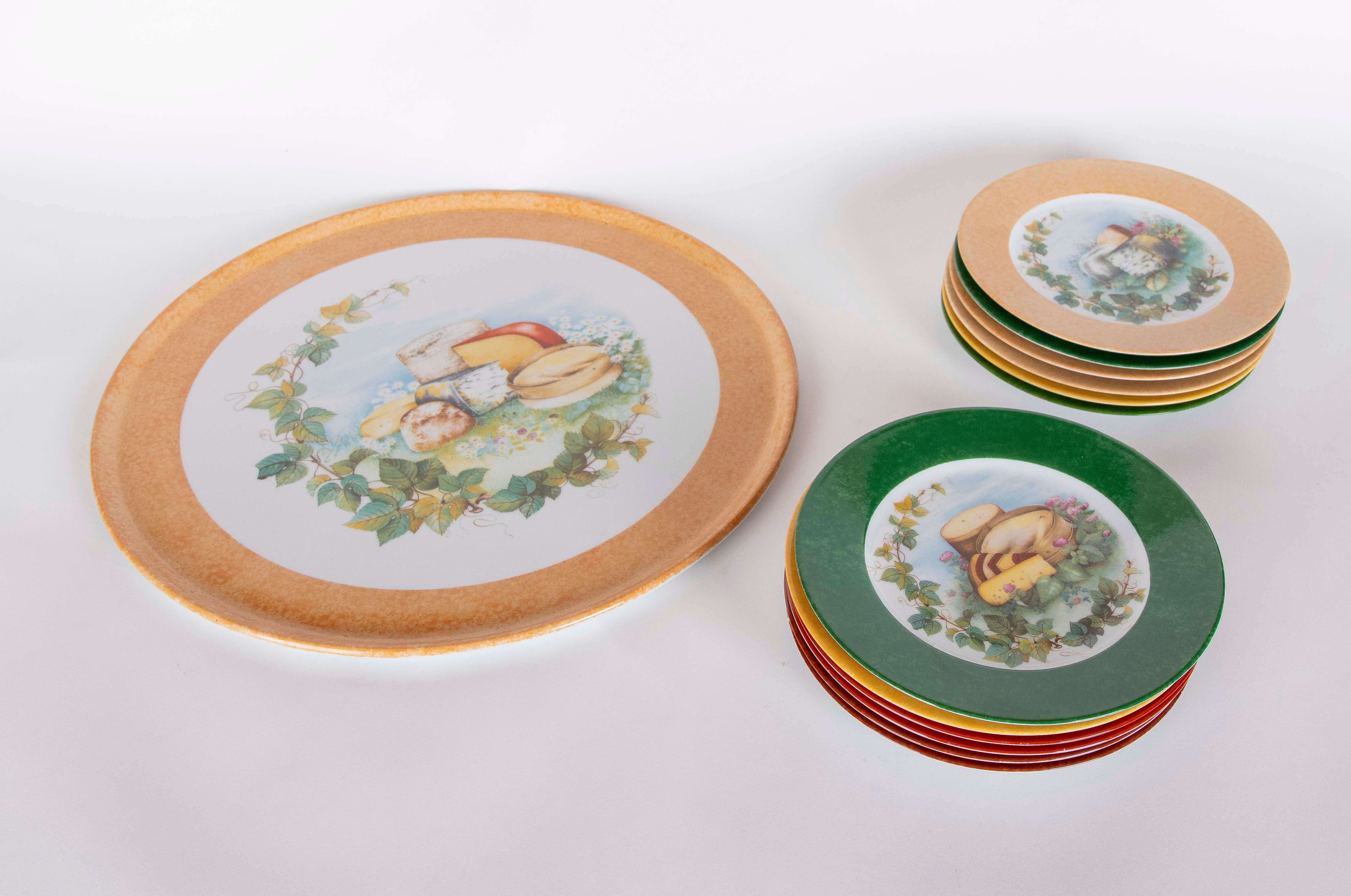 Set of Laure Japy limoges ceramic plates decorated with illustrations of cheese and fruit in a natural setting, surrounded by leaves and flowers. The set includes a large plate with rim in warm tones and several small plates with rims in vibrant