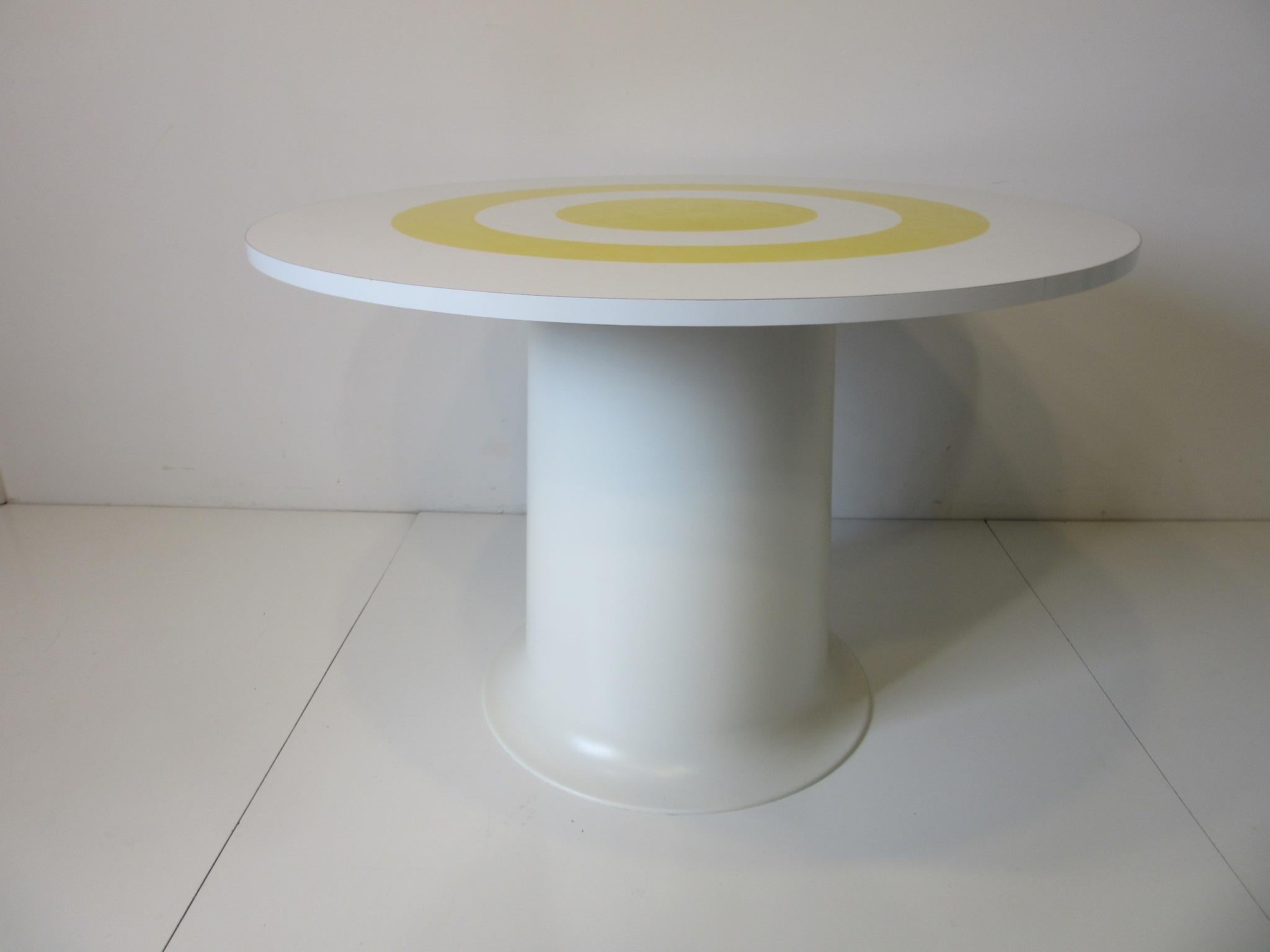 A Space Age dining table with a round laminate top and a bright yellow bulls eye design to the surface sitting on a molded fiberglass and ABS plastic pedestal base in white. Brighten up that small dining area, party room or play room with this fun