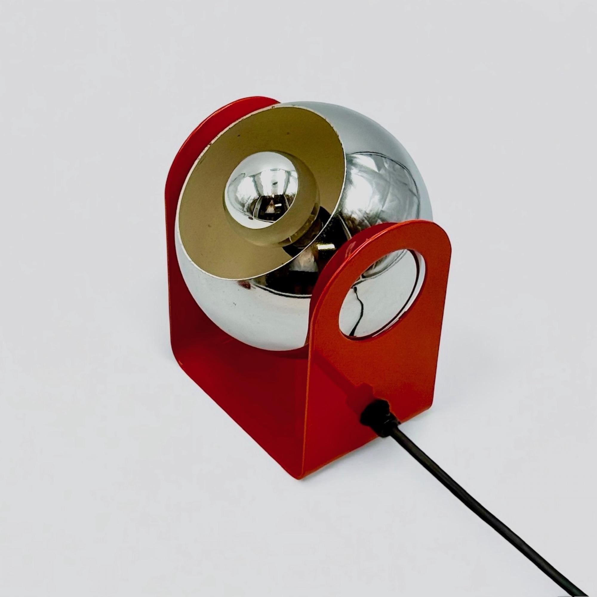 This vintage eyeball lamp is a striking example of Italian Space Age design from the 1970s. Featuring a bold red lacquered U-shaped metal base and a chrome-plated spherical reflector, it embodies the playful yet futuristic aesthetic typical of the