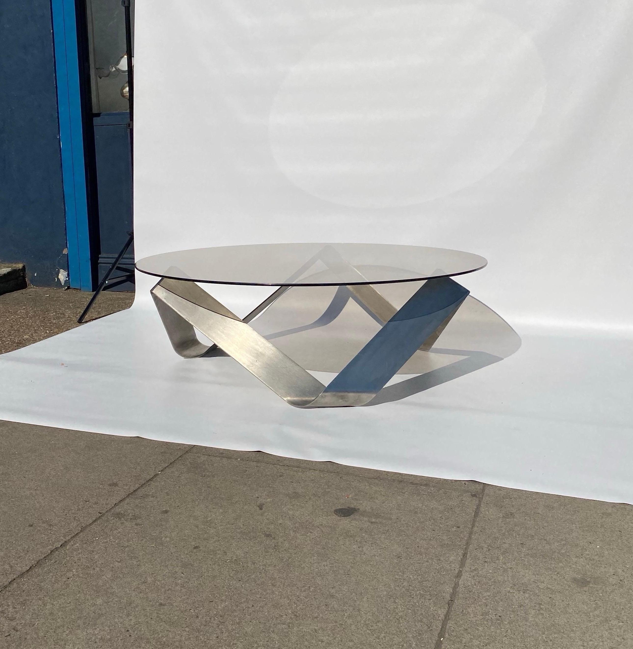 An intricate coffee table by Knut Hesterberg for Ronald Schmitt made in the 1970s.
The frame is made out of brushed aluminium steel following an intricate organic continuous interlocking shape with a round brown smoked glass top. 

With a twist of