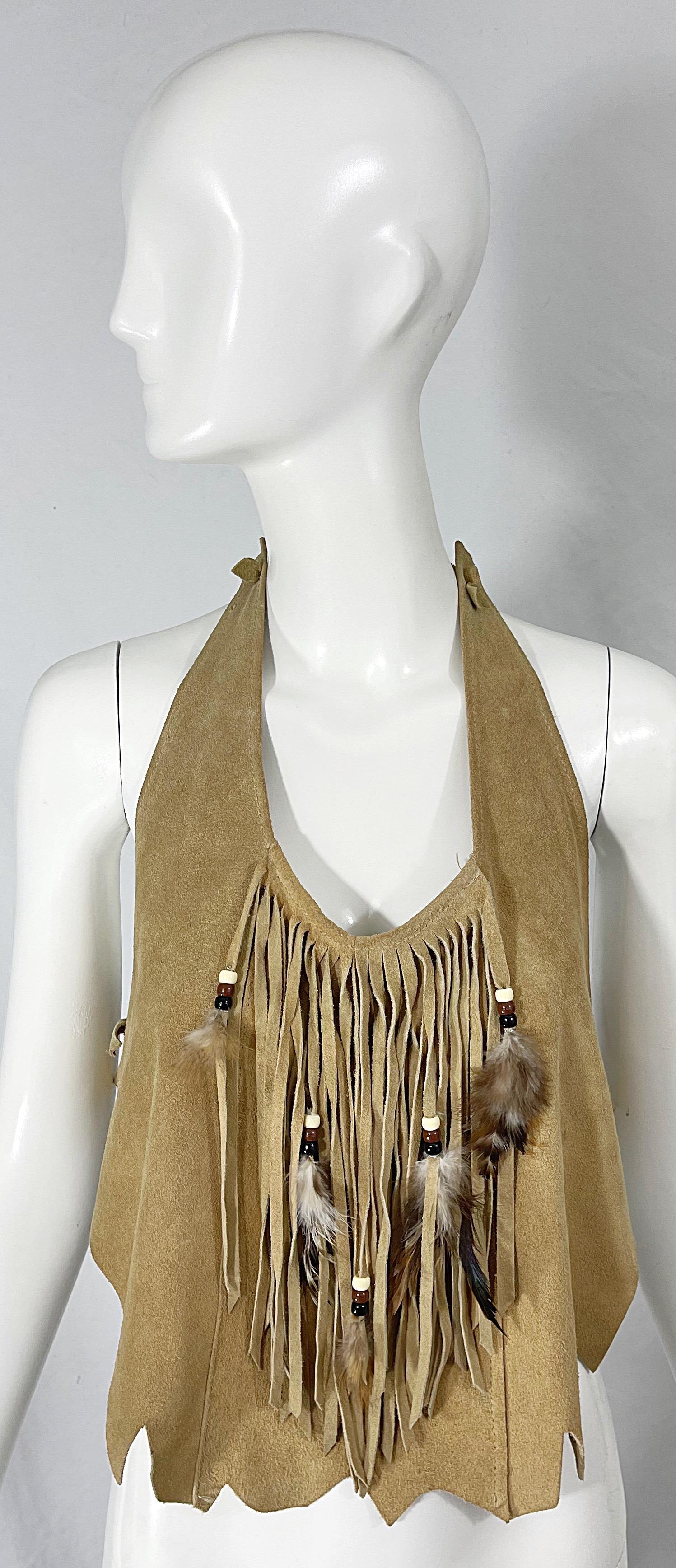 Top boho chic des années 1970 en cuir suédé frangé et perlé ! Il est doté de liens autour de l'encolure et au centre du dos. Franges latérales avec plumes. Ourlet asymétrique. Très flatteur. A associer avec un jean, un short ou une jupe. 
Très bien