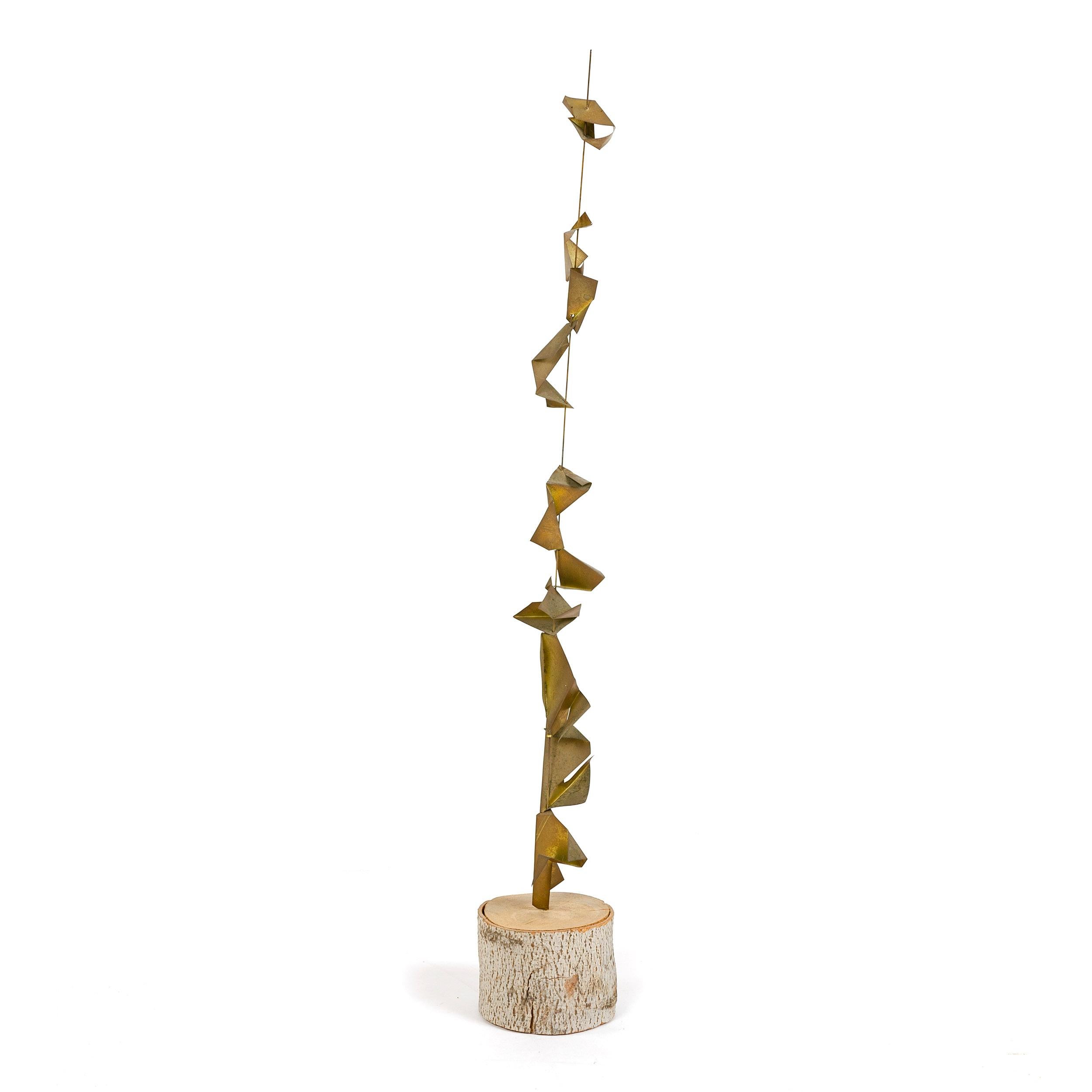 A metal sculpture consisting of a thin rigid wire with folded, fortune-cookie-shape pieces of naturally patinated brass slipped onto the rod and stacked vertically. The rod is set in a round, drum shaped slab of birch retaining its original bark.