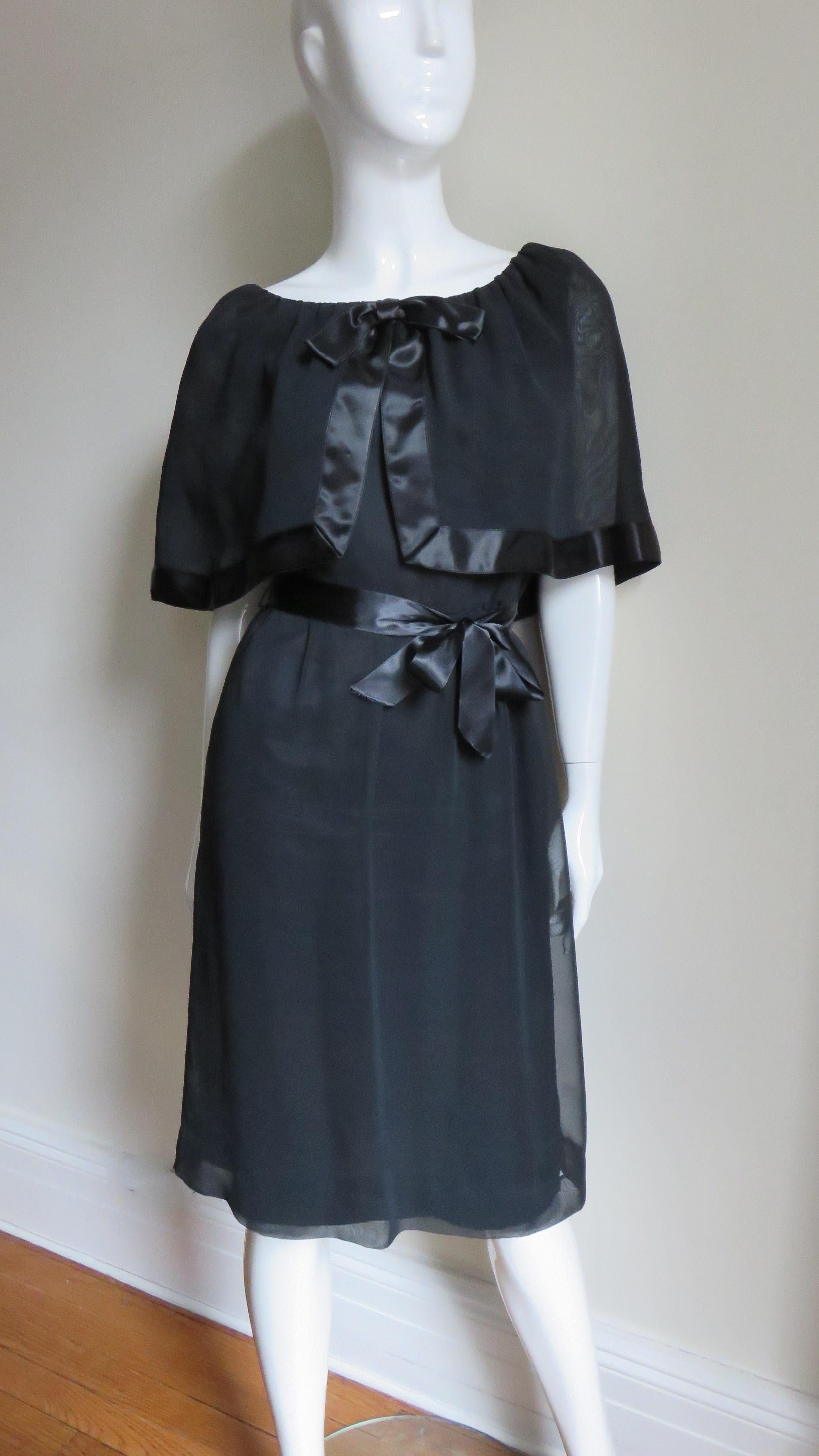 A fabulous black silk dress by William Travilla.  It has a rounded neckline with an attached gathered elbow length caplet edged in black silk satin.  It has a gathered subtle A line skirt, back zipper and a matching black satin tie belt at the
