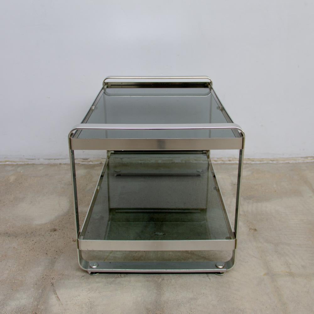 1970s smooth edged two level smoke glass top side table. Primarily used for audio systems or tv. Blends in perfectly in an eclectic scenery. In good vintage condition. Also available a magazine stand in the same style. Measures: 70 x 42 x 43cm.
 
