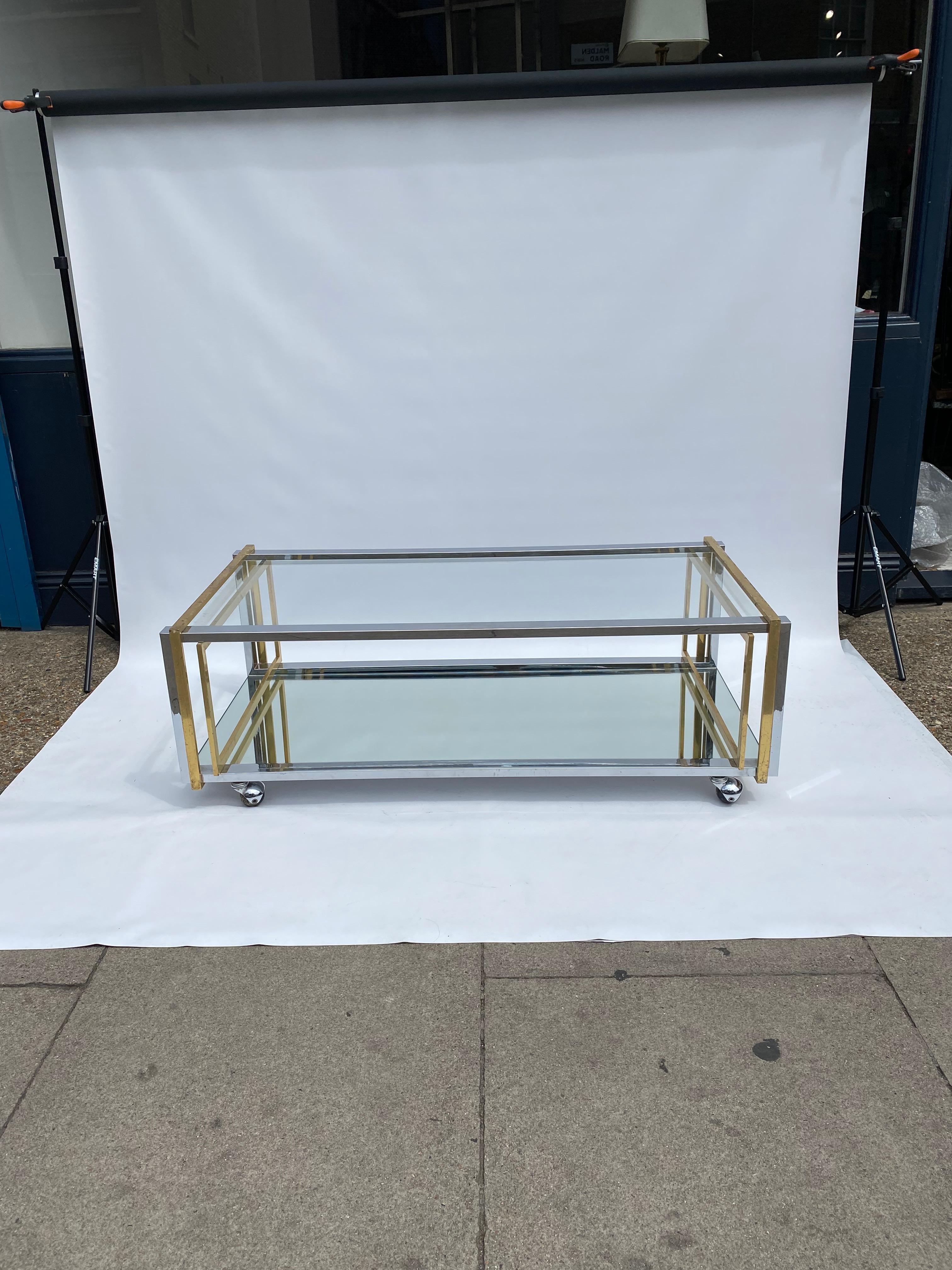 Elegante mesa de centro rectangular de dos niveles con ruedas de la década de 1970, a la manera de Renato Zevi y Romeo Rega y del periodo glam de la Regencia de Hollywood.

El marco de latón y cromo soporta dos niveles de paneles de cristal y