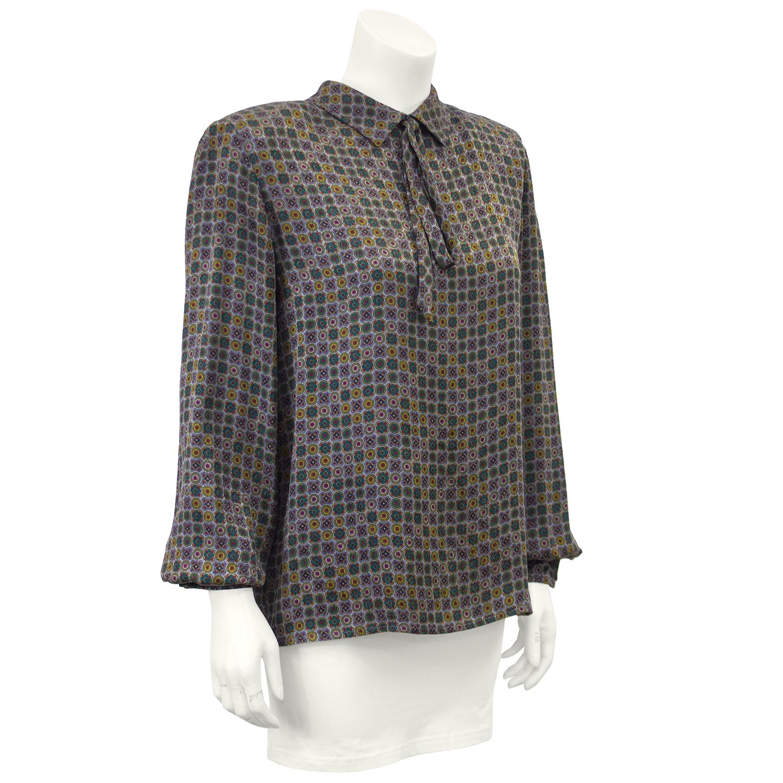 This 1970s Valentino silk blouse features an all over jewel tone pattern on a blue grey background. The petite pointed collar has a fixed loose, long bow at the front and fastens up the back with buttons hidden under a placket and hooks at the nape