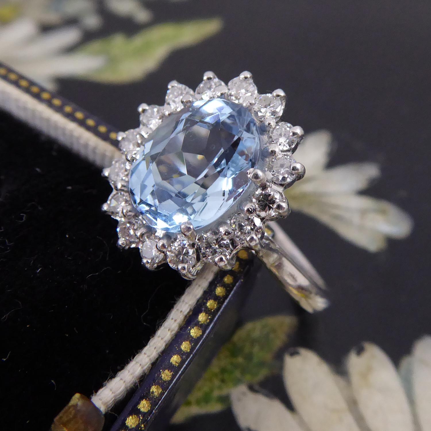 An aquamarine and diamond cluster ring centring on an oval, mixed cut aquamarine of medium colour and measuring approx. 9.54mm x 7.69mm x 5.06mm deep.  The aquamarine is white claw set to a surround of 16 round brilliant cut diamonds measuring