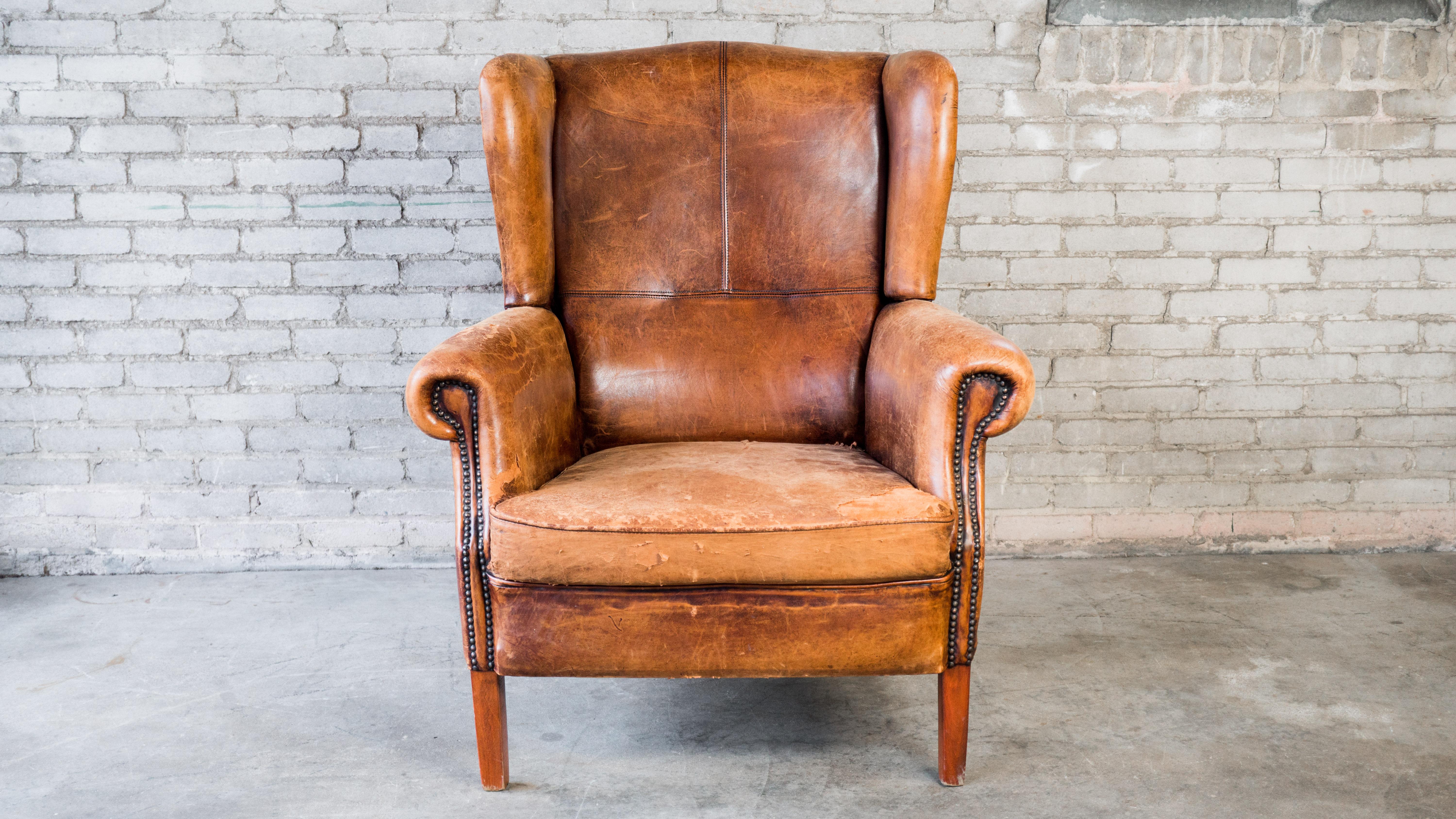 Vintage Art Deco leather wingback chair, circa 1970s. Beautifully distressed cigar brown leather with aged patina. European traditional wingback design with exposed stitching details and brass nailhead trim. Supported by wooden legs. Good vintage