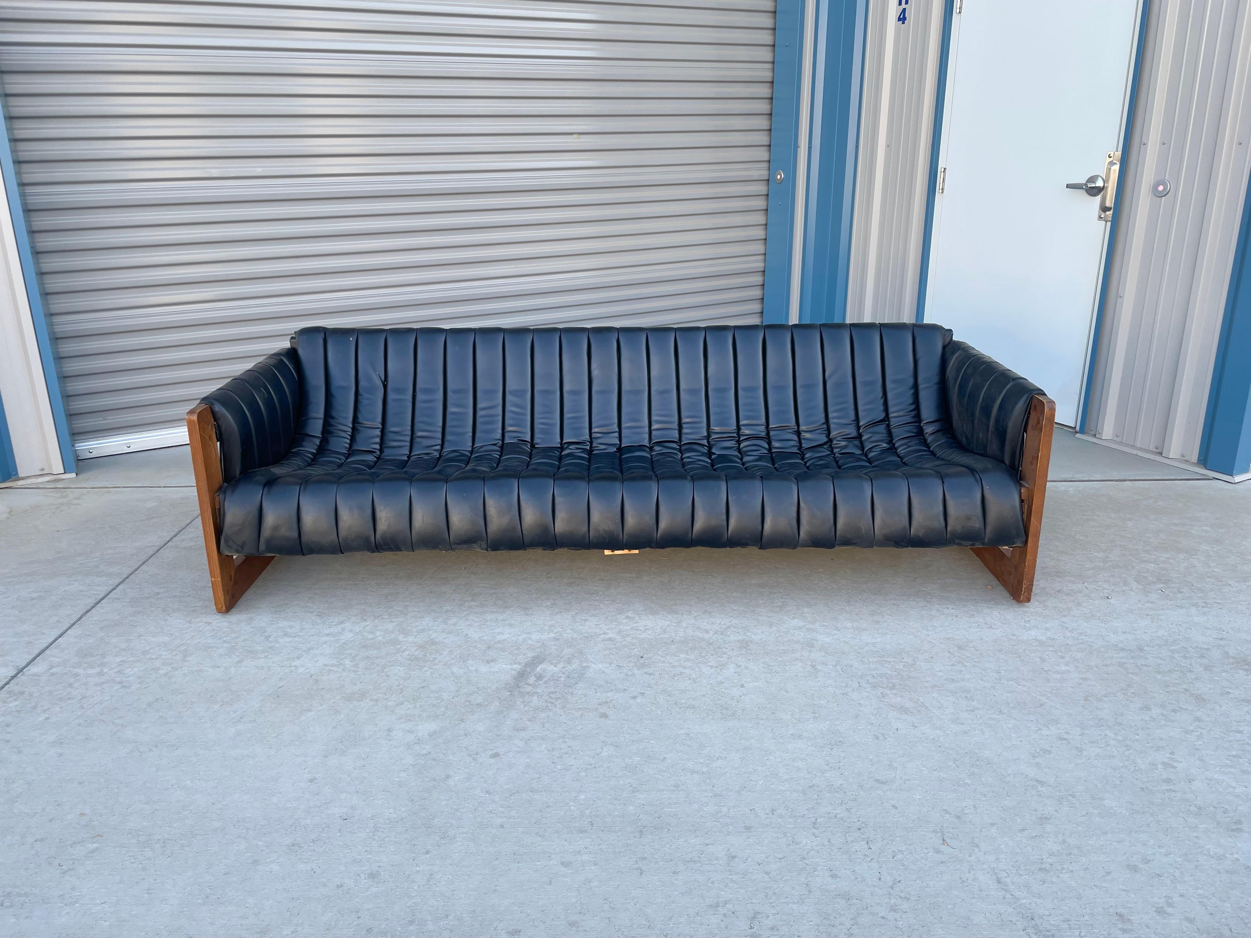 Canapé en cuir noir Hollywood regency conçu et fabriqué aux États-Unis vers les années 1970. Ce magnifique canapé vintage est doté d'une assise en bandoulière touffetée et de pieds uniques en bois dur de noyer, ce qui en fait une pièce unique. Ce