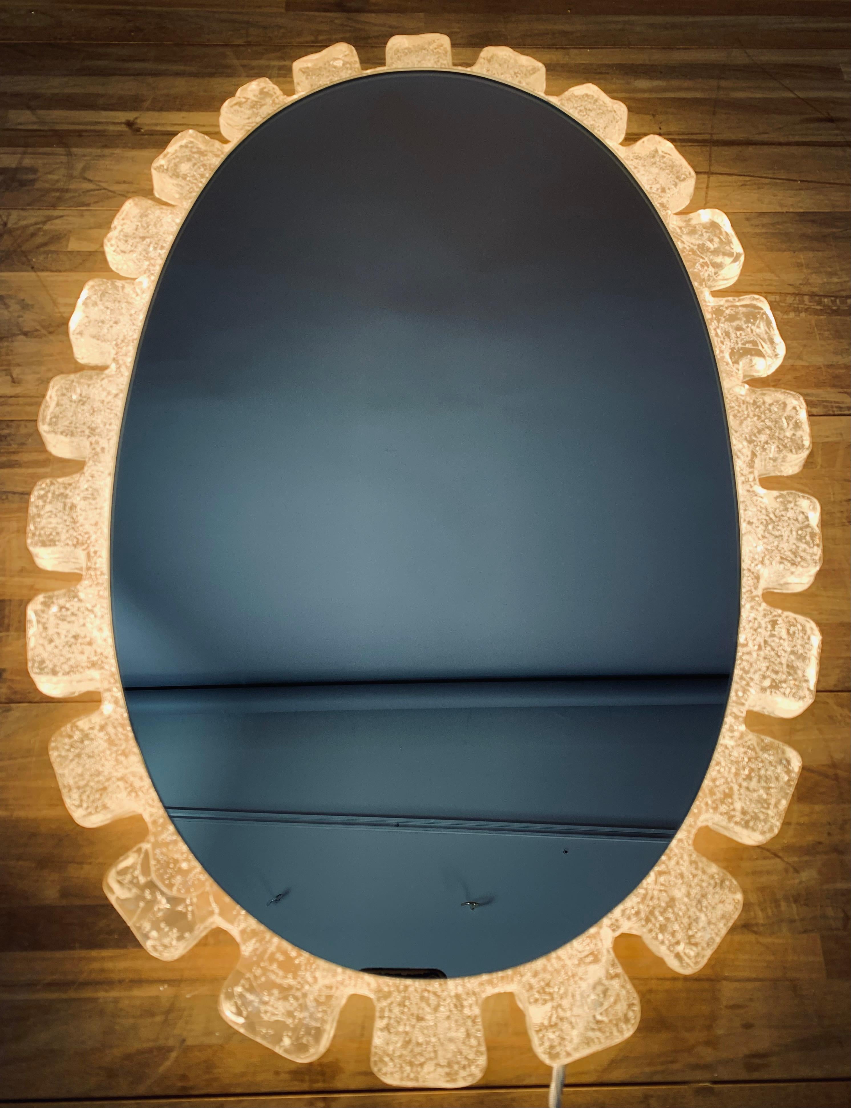 1970s German Hillebrand oval wall mirror with a frosted translucent acrylic frame. The mirror is supported by a white metal frame which the mirror hooks onto and also holds the four lightbulbs. The lucite frame glows evenly when lit and sheds a