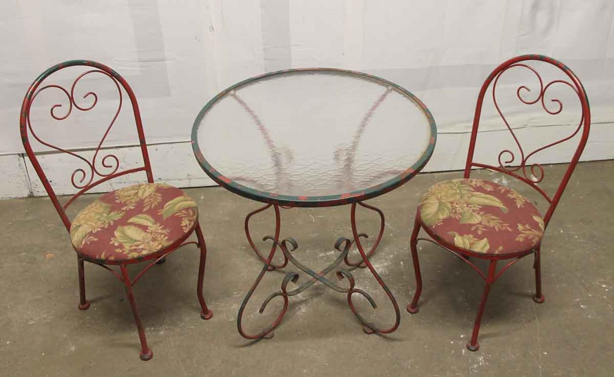 1970s distressed red and green painted steel patio table set. Set includes one round glass top table table and two chairs with leafy floral upholstery. This can be seen at our 400 Gilligan St location in Scranton, PA. 