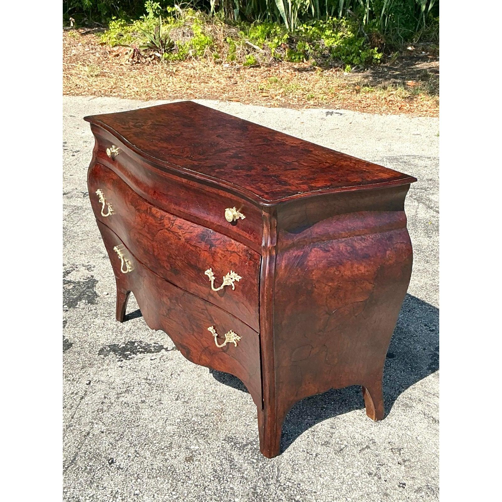 Diese exquisite Bombenkommode im Vintage-Stil ist ein wahres Fest der klassischen Regency-Eleganz, mit einer reich gemaserten Oberfläche aus olivfarbenem Wurzelholz und einer anmutigen Serpentinen-Silhouette. Die verschnörkelten Messingbeschläge