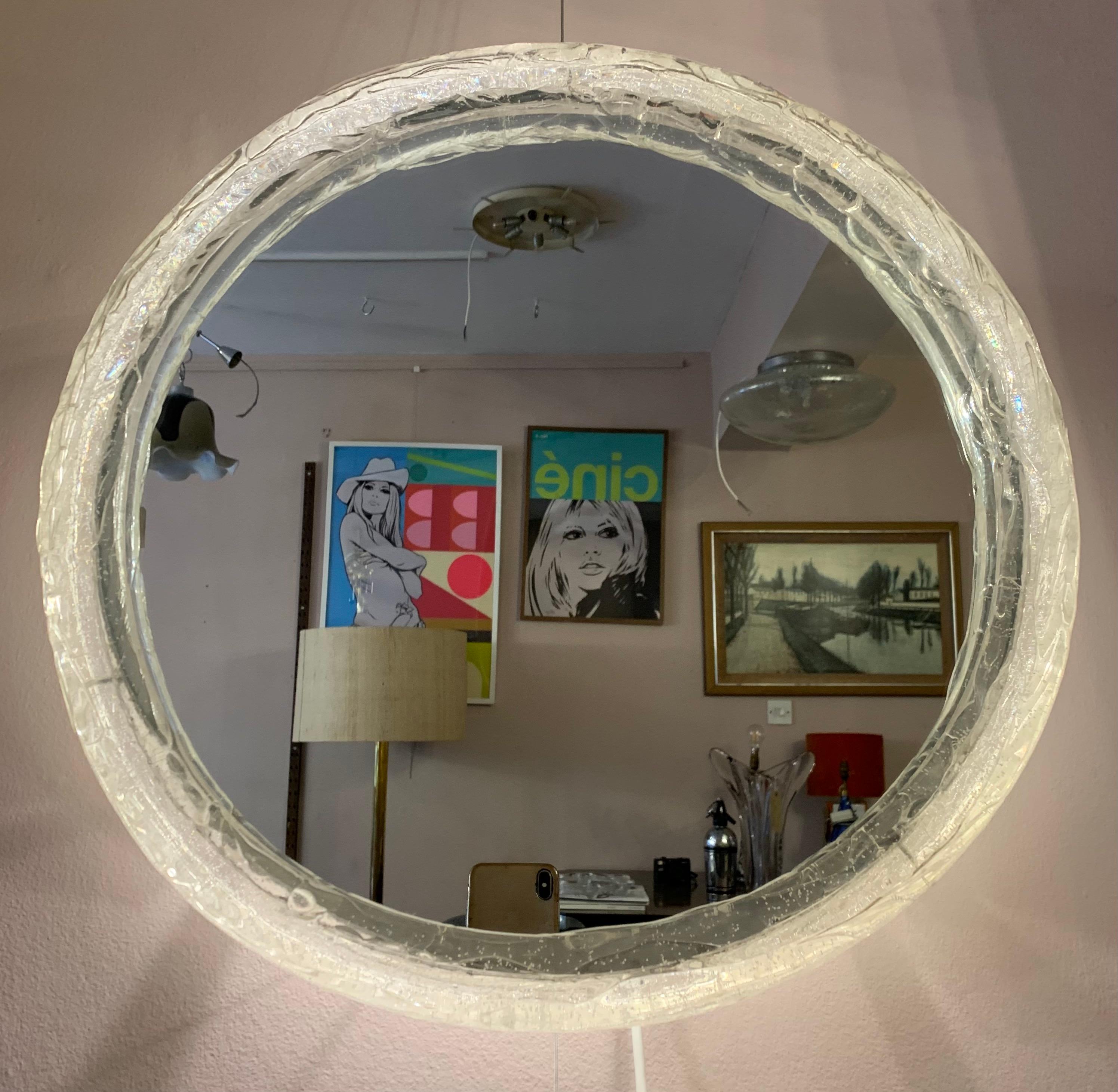 A small 1970s German illuminated, iced lucite-framed, circular, mirrored-glass wall mirror.   The glass mirror is surrounded by a lucite round frame which the light shines evenly through via the three evenly spaced bulbs.  The mirror and frame hook