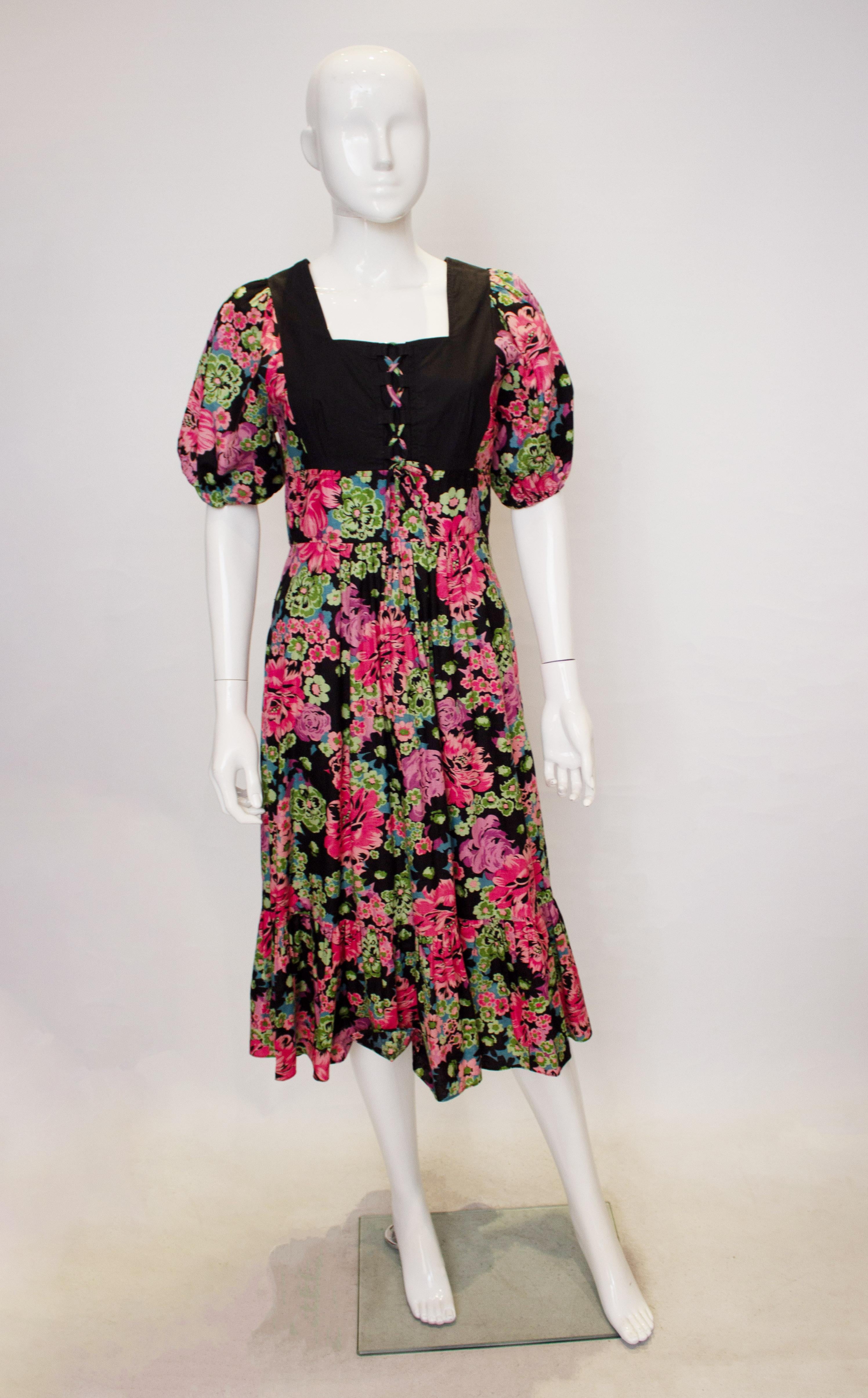 Ein tolles Vintage-Kleid für den Sommer von Susie G. Das Kleid hat einen quadratischen Ausschnitt und Rückenausschnitt, mit leicht gepufften Ärmeln. Es hat eine zentrale Öffnung am Rücken, Rüschen am Saum und eine Schnürung am Mieder.