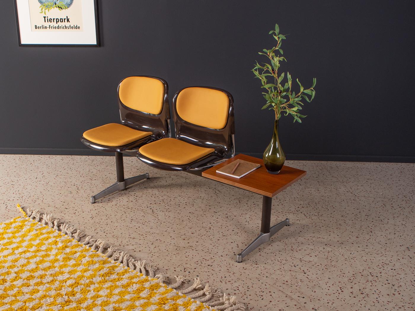 Classic waiting bench from the 1970s. High-quality steel and stainless steel frame with two plastic seat shells in brown and a table top in teak veneer. The seat shells have been reupholstered and covered with a high-quality leatherette in ochre.