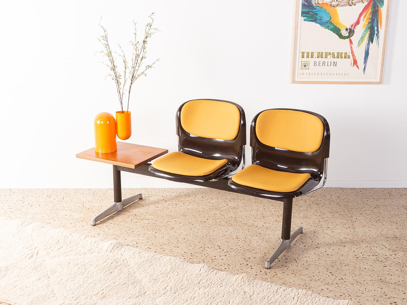 Classic waiting bench from the 1970s. High-quality steel and stainless steel frame with two plastic seat shells in brown and a table top in teak veneer. The seat shells have been reupholstered and covered with a high-quality leatherette in ochre.