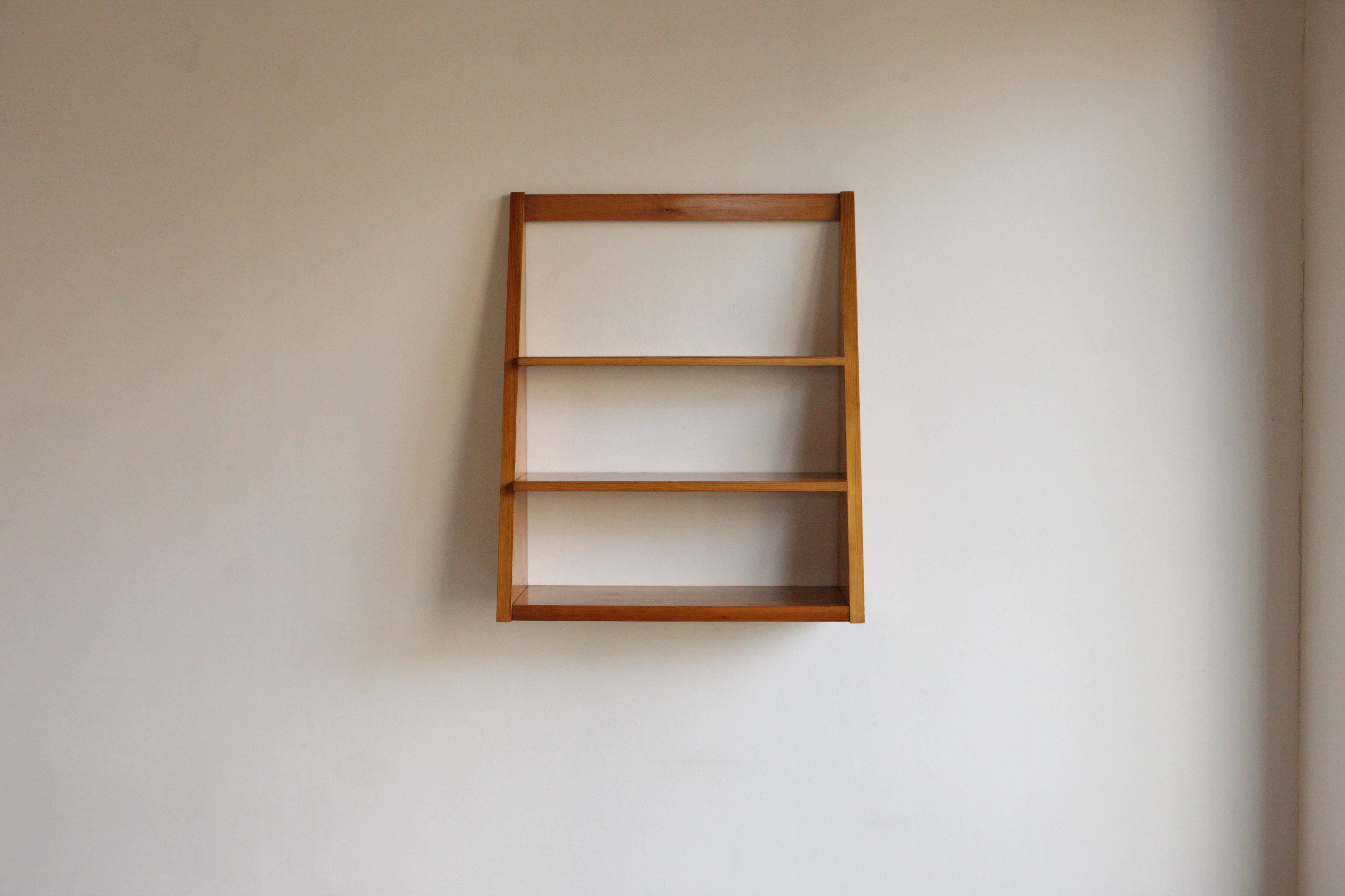 A Perriand style wall hanging shelf in solid pine with a heavy grain. Three shelves of staggered depth sit within a frame that fixes onto the wall. Circa 1970's.