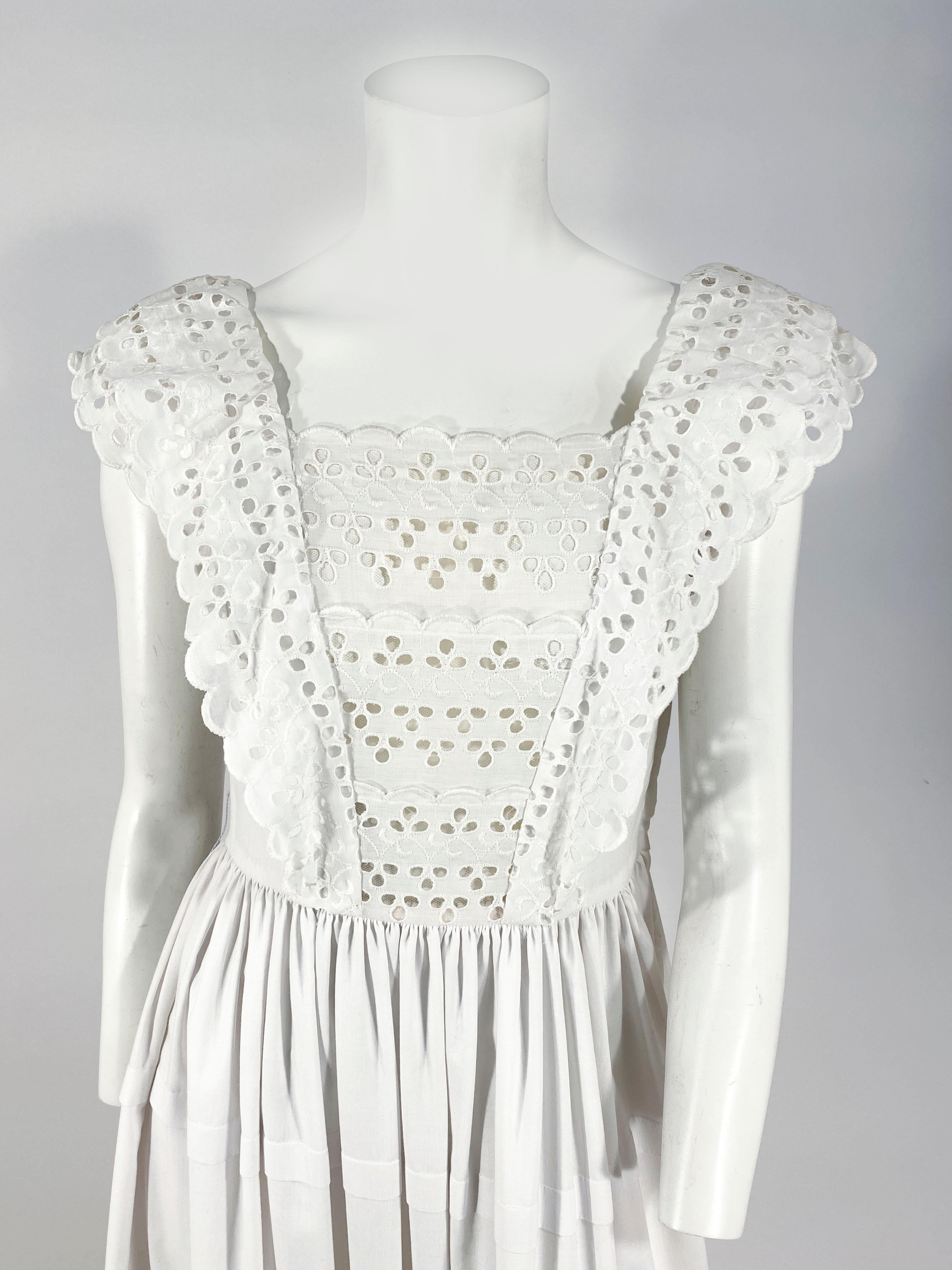 1970s white cotton cottage dress with eyelet lace ruffles along an applied pinafore silhouette and up over the shoulders. The skirt has a wide layered ruffle hem and tiers of pleats. The bodice has a hidden side zipper closure. 