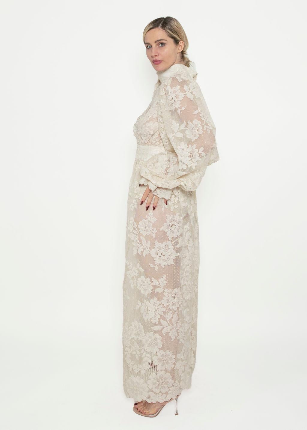 Our stunning 1970's white lace gown features a beautiful full floral lace fabric and a mock neck design.  It is complete with a keyhole chest and balloon sleeves which have elastic at its cuffs. A unique open keyhole back adds a touch of elegance