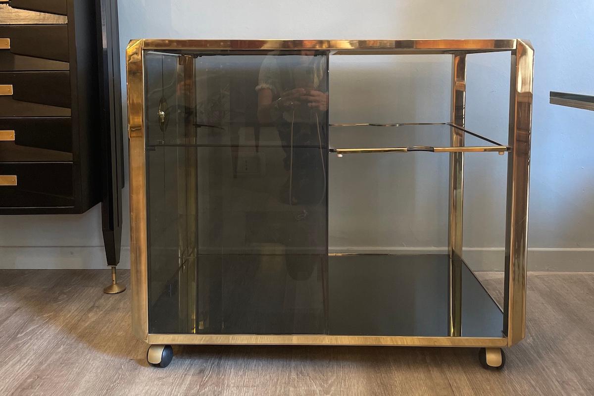 1970s bar cart designed by Willy Rizzo, made of brass and smoked glass. 
The piece is divided into two sections: an open area featuring a practical pull-out shelf that serves as a tray, and a closed section with two glass doors and brass handles,