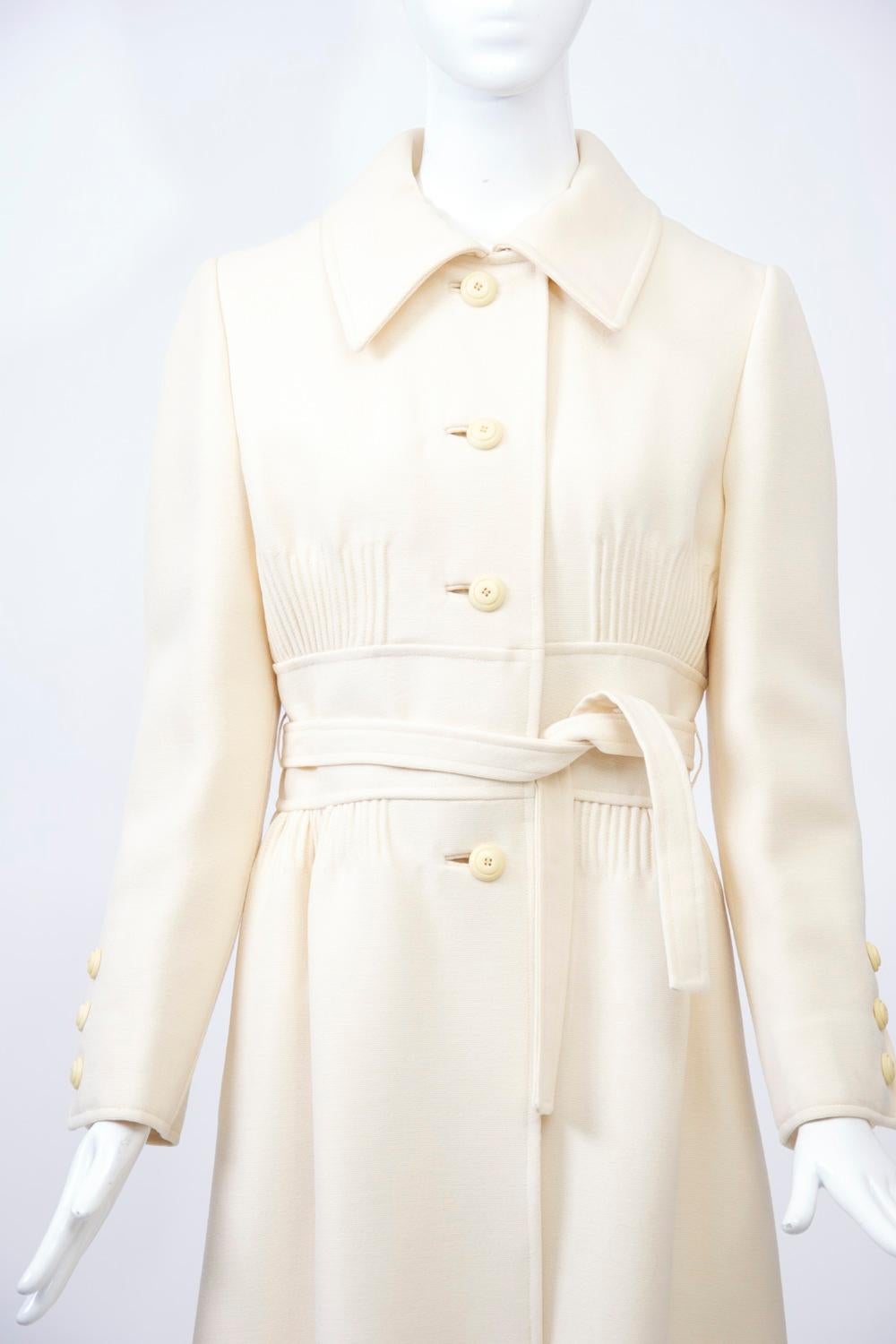 Beautifully detailed 1970s coat in off-white wool. Unusual features include a wide inset 
welted midriff band with tucking above and below in front finished with a self-tie belt. Single breasted with with shirt collar and ivory colored buttons in